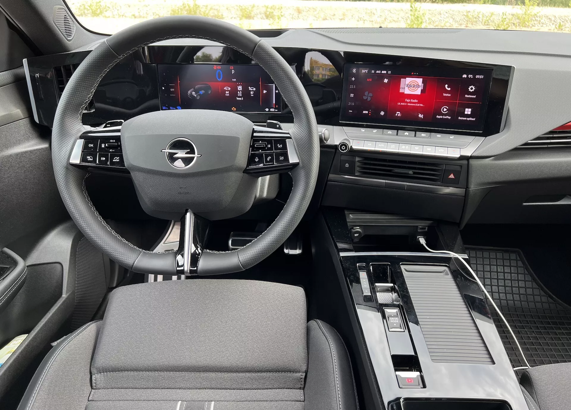 Vista interior do Opel Astra 2022 a partir do banco traseiro, mostrando o volante de três raios com controles integrados, cluster de instrumentos digital, tela touchscreen de infotainment, console central com alavanca de câmbio e armazenamento, e uma mistura de materiais em tecido e efeito de metal