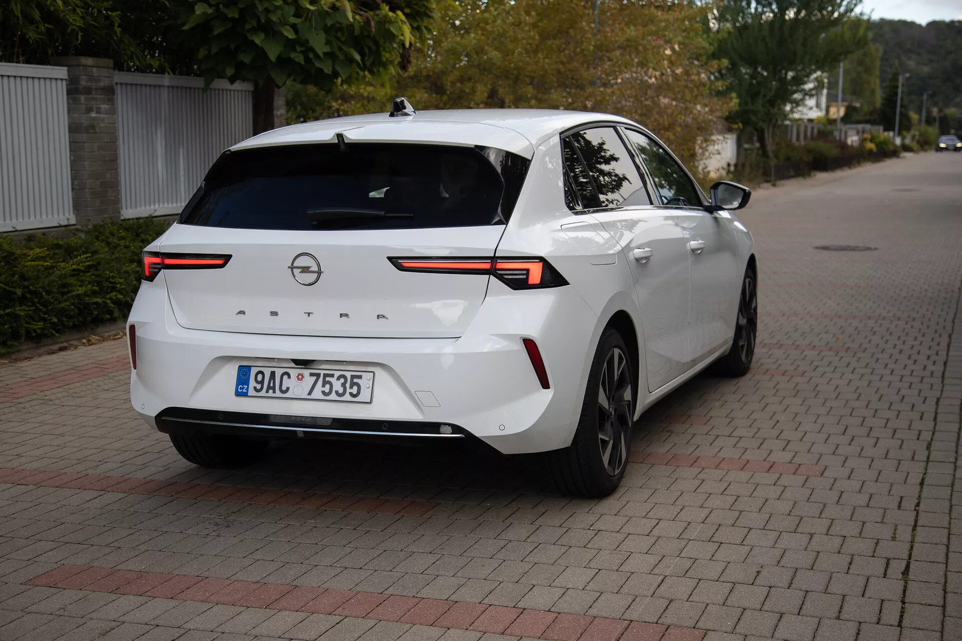 Sedã moderno Opel Astra 2022 em branco, estacionado sobre superfície de tijolos interligados, exibindo ângulo traseiro de três quartos com foco em luzes traseiras LED de alta tecnologia, câmera de ré, sensores de estacionamento, perfil lateral parcial incluindo roda traseira direita e porta, conectados por uma barra de luz horizontal através da tampa do porta-malas, com antena em forma de barbatana de tubarão para conectividade aprimorada, adequado para ilustrar o design traseiro do veículo e tecnologia automotiva moderna