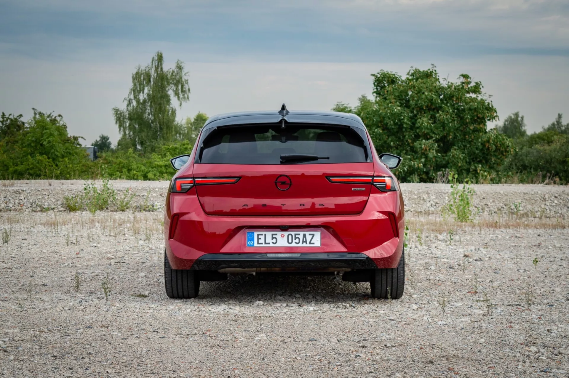 Opel Astra 2022 fotografado de trás em uma superfície de cascalho exibindo lanternas traseiras com uma barra de luz conectando, sensores de estacionamento e ausência de portas de escape indicando um possível modelo elétrico ou híbrido, contra um pano de fundo de vegetação e céu ligeiramente nublado.