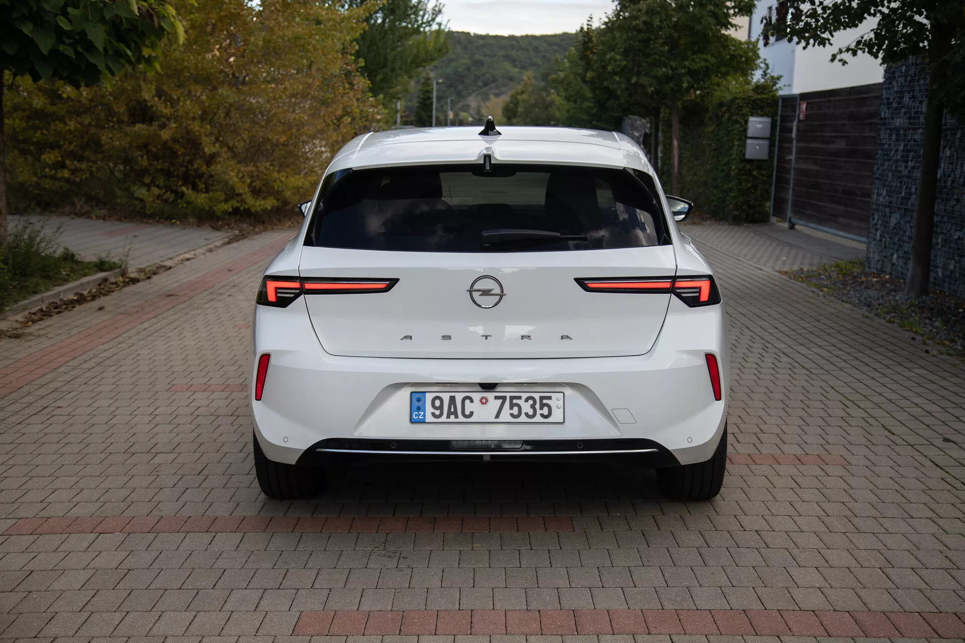Vista traseira de um Opel Astra 2022 branco exibindo luzes traseiras LED, sensores/câmera de estacionamento traseiro, emblema da marca, placa de licença, aerofólio traseiro, luz de freio alta, antena em forma de barbatana de tubarão e acabamento de pintura limpo, estacionado em uma superfície com padrão de tijolos à luz do dia.