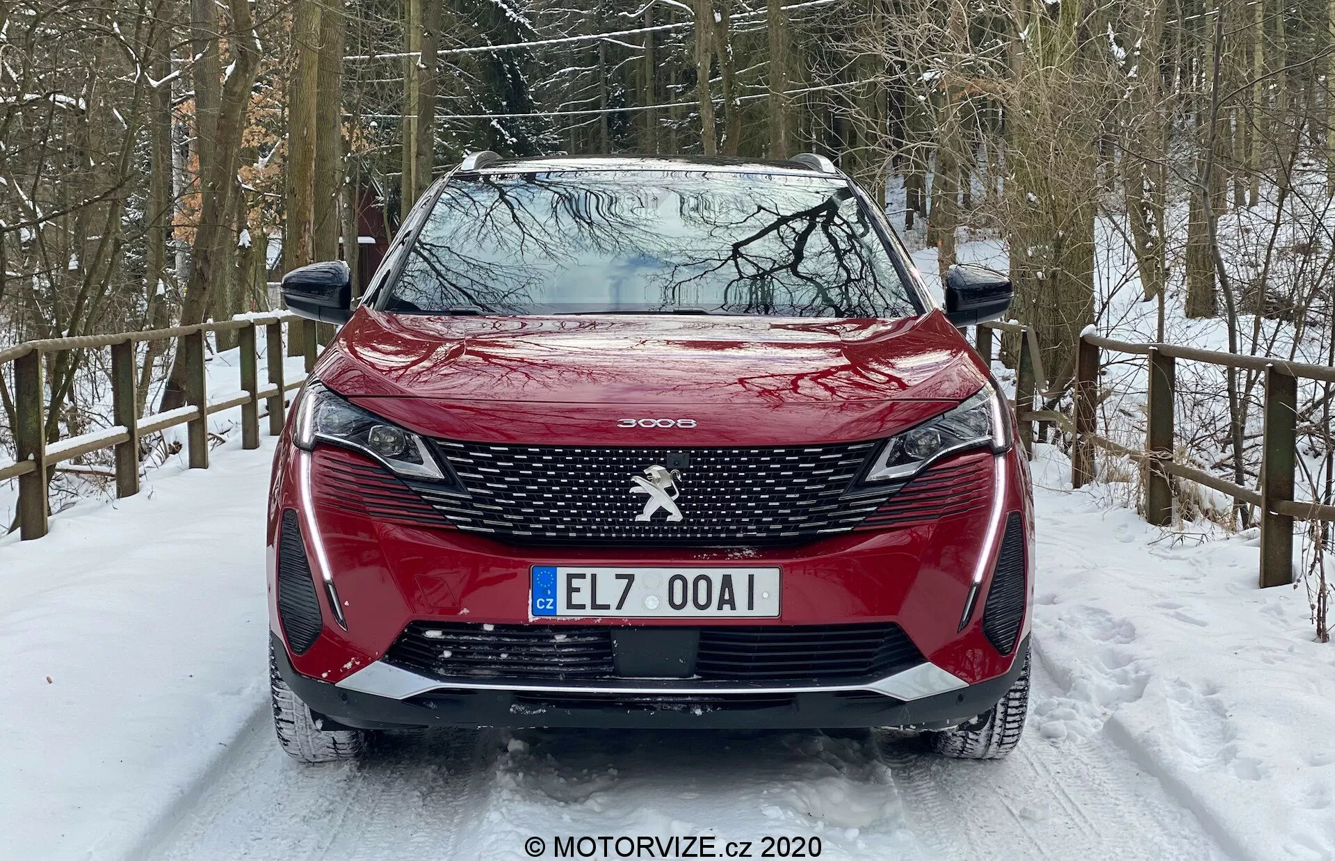 Vista frontal de um Peugeot 3008 vermelho 2021 estacionado numa estrada coberta de neve, com padrão de grelha distintivo, emblema central da Peugeot, faróis dianteiros afiados com DRLs tipo presas, capô contornado, para-choque dianteiro com sensores de estacionamento integrados e um ambiente florestal nevado.