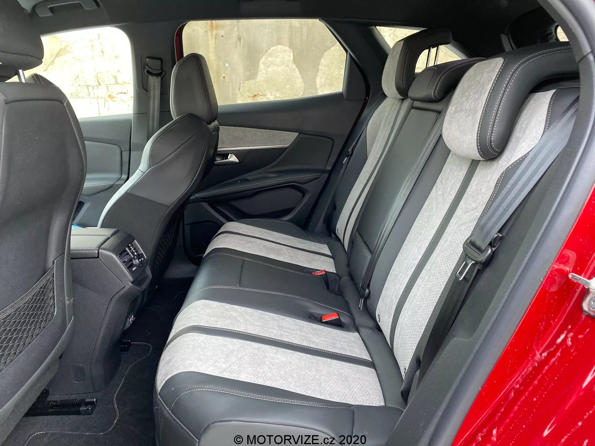 Foto do interior de um Peugeot 3008 de 2021 focando na área dos assentos traseiros visto de fora, inclinado para o lado do motorista. Os bancos traseiros apresentam um tecido bicolor com tons de cinza escuro e claro e um padrão de listras texturizado. Os apoios de cabeça levantados correspondentes são visíveis. Parte do assento da frente e do console central também estão à vista. Os bancos traseiros têm cintos de segurança de três pontos e um apoio de braço com porta-copos integrados. Os painéis das portas correspondem às cores dos bancos com grades de alto-falantes e seções de controle de energia.