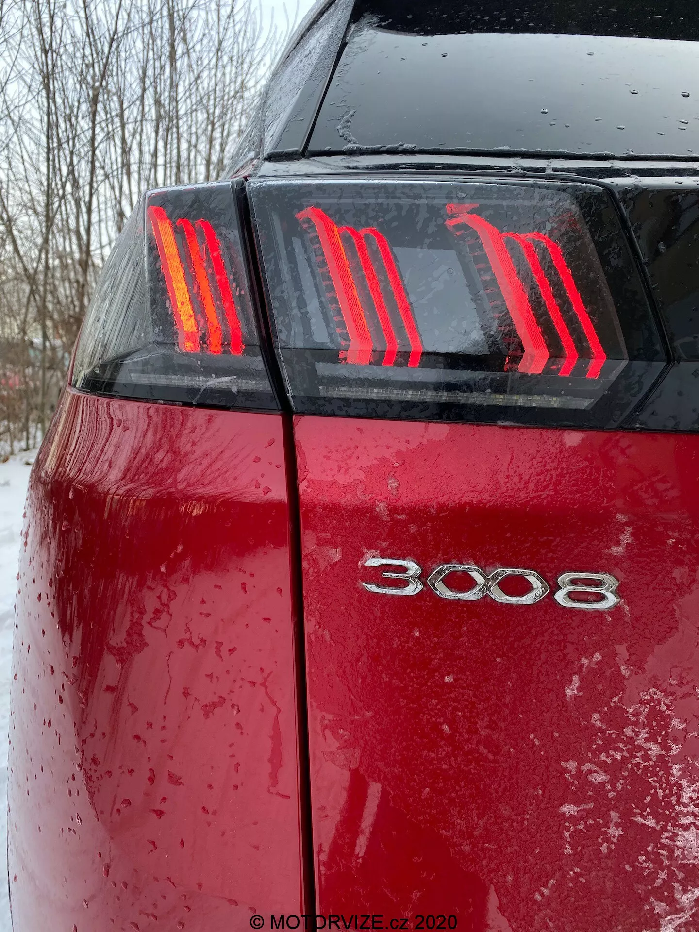 Close-up da porção traseira do Peugeot 3008 2021 vermelho a partir de um ângulo baixo, mostrando as distintas luzes traseiras LED com três tiras verticais em forma de garra e a insígnia do modelo '3008'. A superfície molhada do carro, coberta de gotas de água e sujeira ou neve, destaca o uso recente em condições meteorológicas adversas com condições de iluminação nubladas.