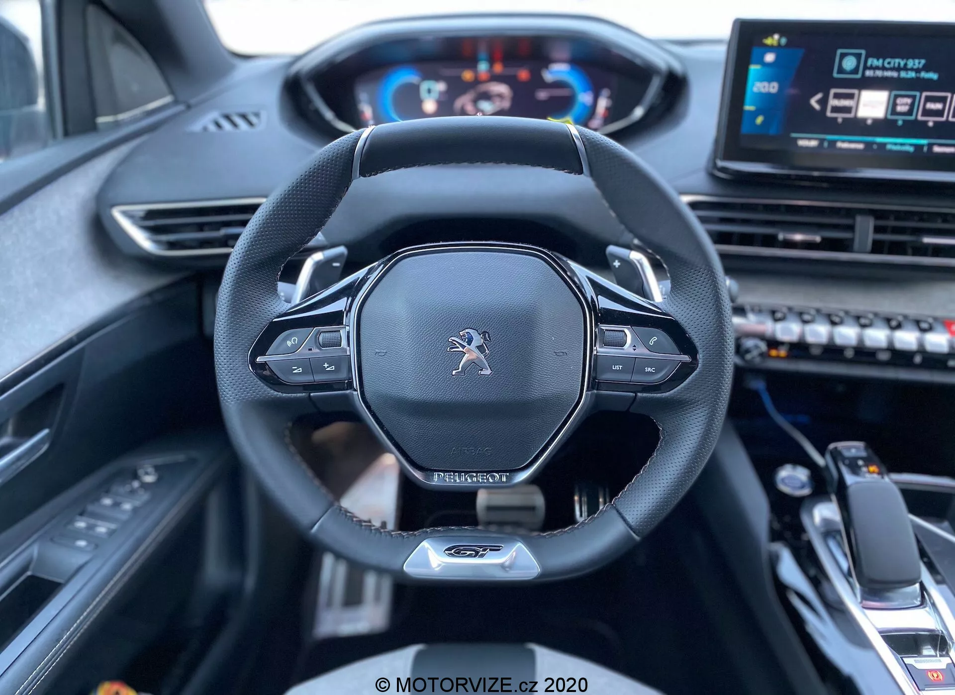 Vista interior do banco do motorista de um Peugeot 3008 2021 mostrando o volante e o painel. A imagem foca-se no volante centralizado com logotipo Peugeot, design moderno com fundo plano, aro revestido em couro e controles montados, provavelmente para configurações de áudio e controle de cruzeiro. As pás para troca de marcha manual são visíveis atrás do volante. Um painel de instrumentos digital exibe o velocímetro e o tacômetro iluminados, indicando que o veículo está ligado. À direita do volante, um sistema central de infotainment com tela sensível ao toque e vários botões de controle são parcialmente visíveis, juntamente com um seletor de marchas e apoio de braço central em primeiro plano inferior, sugerindo um veículo moderno e tecnologicamente avançado com um interior sofisticado.