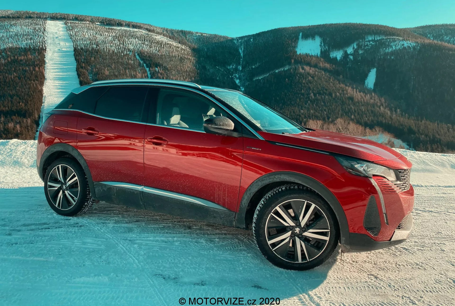 SUV Peugeot 3008 2021 em vermelho, capturado de uma vista frontal de três quartos em uma superfície nevada com um cenário montanhoso, destacando os faróis avançados de LED, rodas de liga complexas, acabamento brilhante, grelhas ativas e retrovisores laterais com piscas integrados, enfatizando os recursos de design e a adequação para condições desafiadoras.