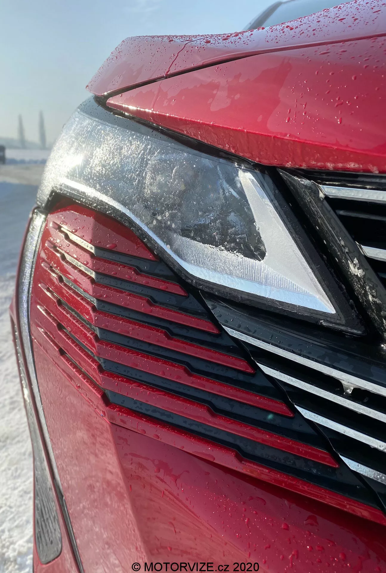 Vista aproximada da parte dianteira do Peugeot 3008 2021, focando no farol LED moderno e na grade com lâminas verticais em um exterior vermelho metálico coberto com gotas de água, sugerindo chuva recente ou geada. O fundo nevado sugere condições de inverno ou detalhamento de cuidados com o carro.