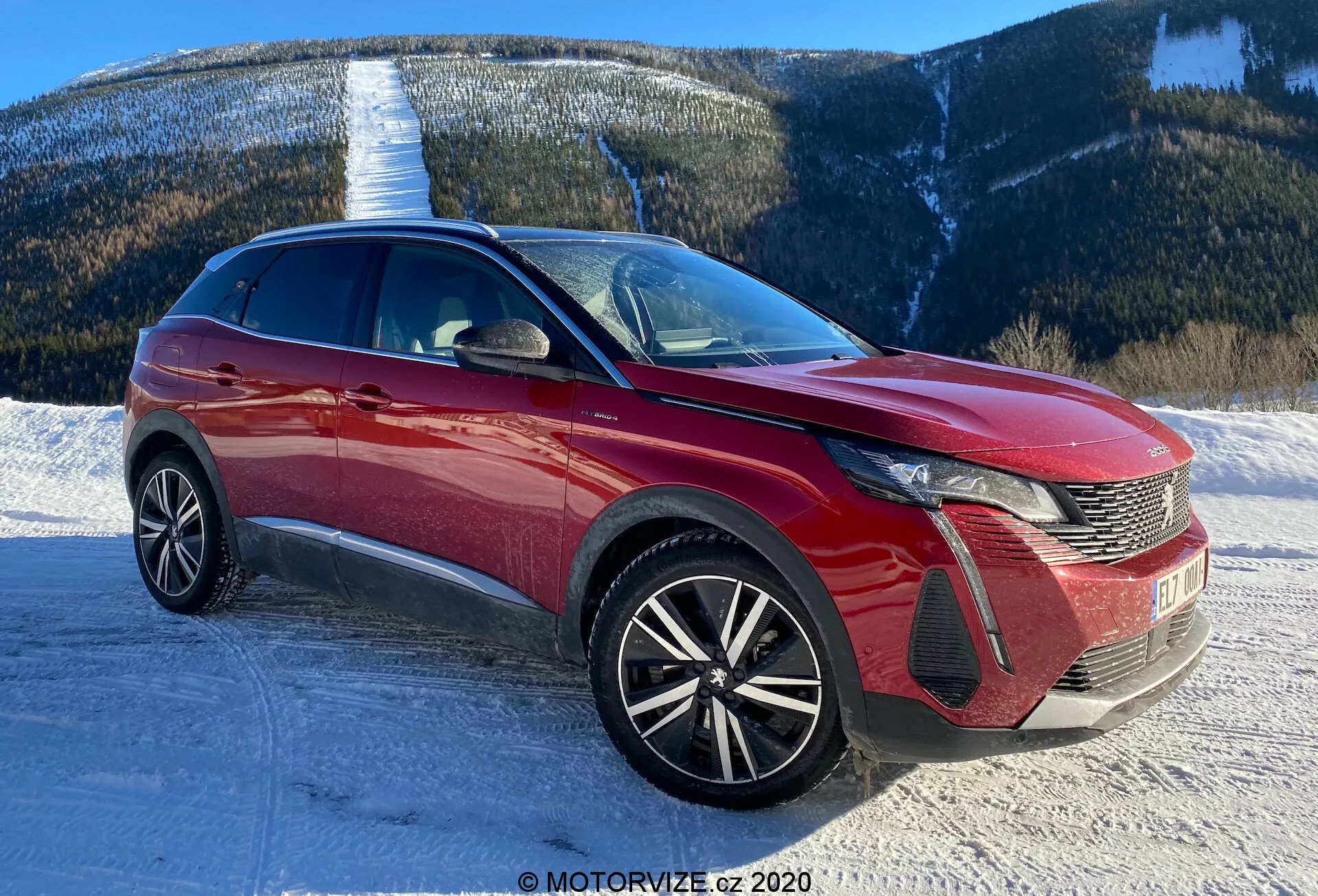 Imagem de um carro moderno, provavelmente um Peugeot 3008 2021, tirada de uma perspectiva de três quartos frontal. O ângulo destaca o design distinto dos faróis LED do carro, a grade com padrão de pente e o emblema na frente. O para-choque dianteiro exibe seções de entrada de ar e alojamentos para faróis de neblina. O capô apresenta linhas esculpidas, contribuindo para a aerodinâmica, enquanto o para-brisa expansivo indica boa visibilidade frontal. As rodas de liga leve multirraios sugerem um nível de acabamento mais esportivo e superior. A linha de teto elegante e as linhas dinâmicas da carroceria indicam um crossover ou SUV. Espelhos laterais com indicadores de direção integrados e elementos de proteção ou detalhes de design são visíveis. O cenário é uma estrada coberta de neve com um fundo de montanha, implicando capacidade de enfrentar diversos terrenos.
