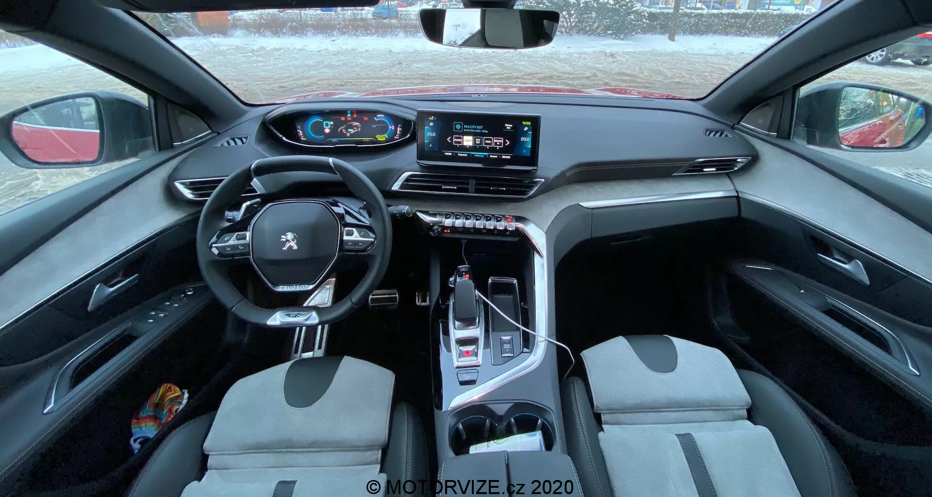 Vista interior do Peugeot 3008 de 2021, mostrando um volante multifuncional revestido em couro com controles montados, um painel de instrumentos digital, um grande sistema de infotainment com tela sensível ao toque na consola central, um seletor de marchas eletrônico, bancos dianteiros com design bicolor e uma mistura de materiais práticos e luxuosos. A foto foi tirada do banco traseiro, oferecendo uma visão abrangente da cabine, destacando o design e os recursos tecnológicos.