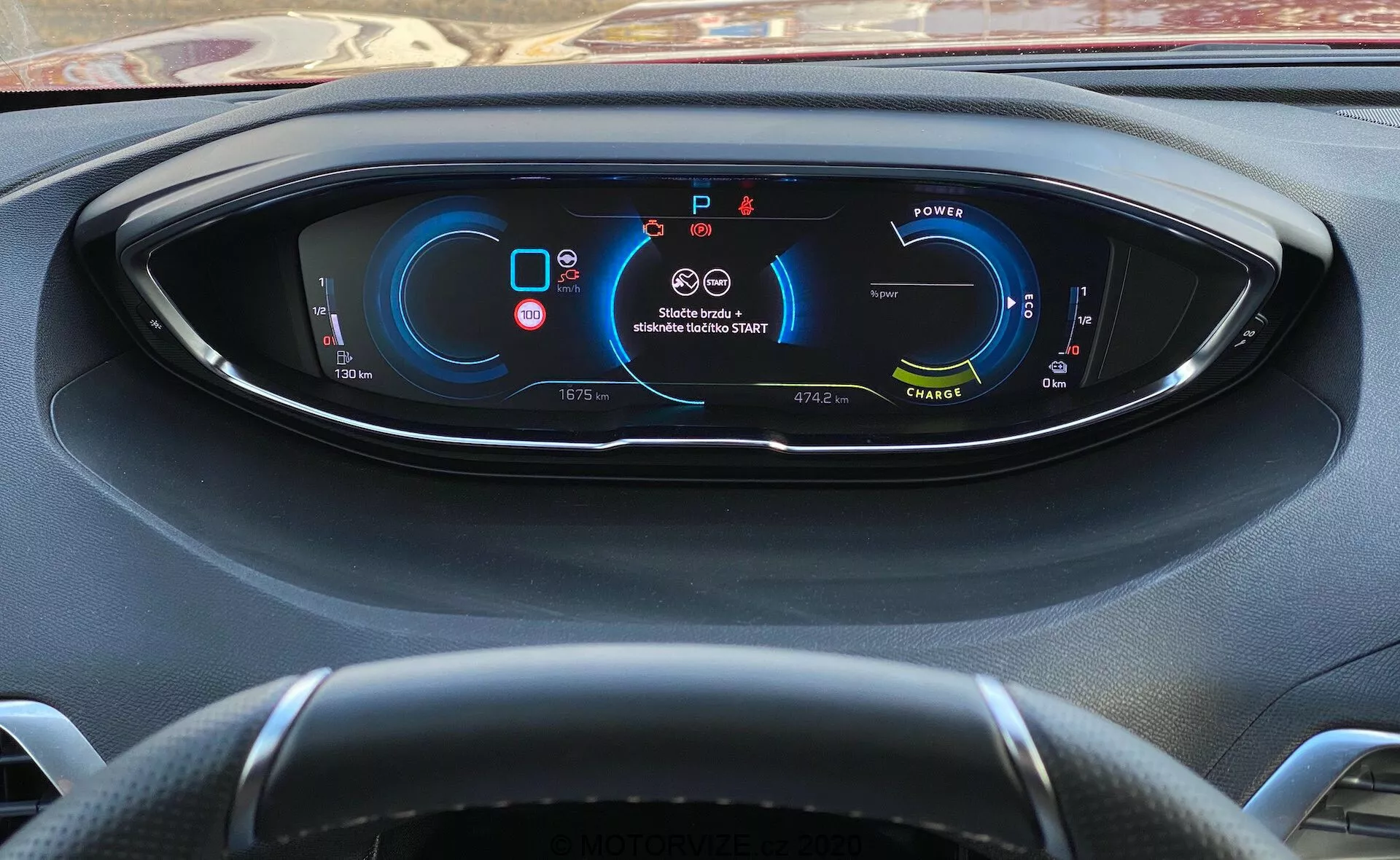 Visão do motorista do painel de instrumentos do Peugeot 3008 de 2021 com painel totalmente digital exibindo métricas e indicadores do veículo. À esquerda, há um medidor de potência, sugerindo um veículo elétrico/híbrido; no centro superior, avisos de cinto de segurança e airbag; à direita, velocímetro indicando que o veículo está parado; na parte inferior, indicações de marcha (estacionado), odômetro, temperatura externa, modos de condução e alcance disponível. Volante de três raios parcialmente visível. Vista desde trás do volante com um leve ângulo para baixo.
