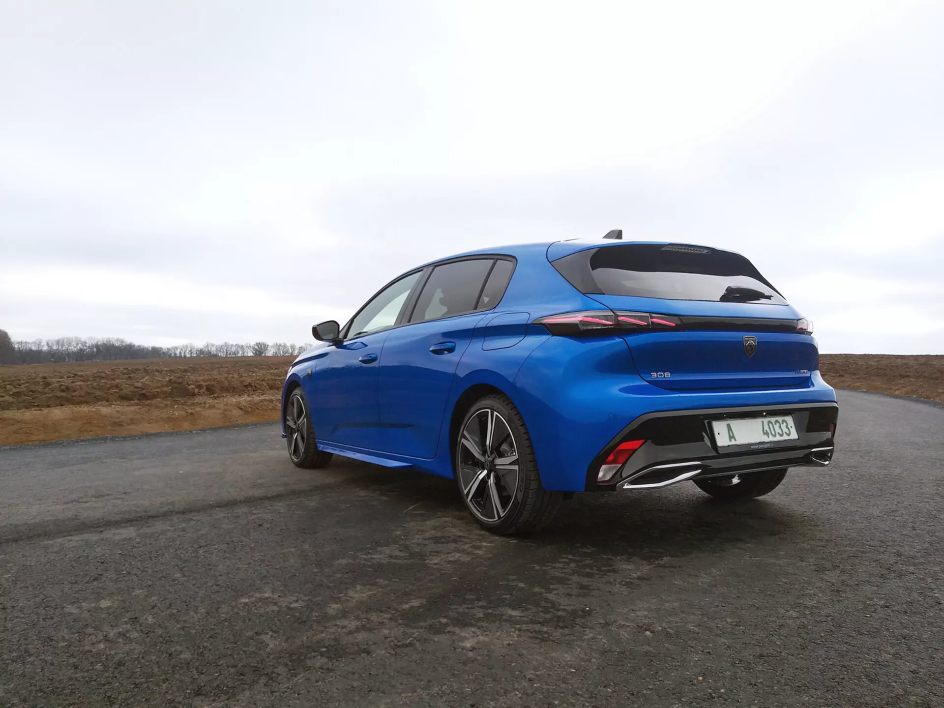 Hatchback Peugeot 308 2022 em azul vibrante, vista traseira de três quartos destacando as luzes traseiras LED, spoiler integrado e rodas de liga leve multirradiadas contra um céu nublado.