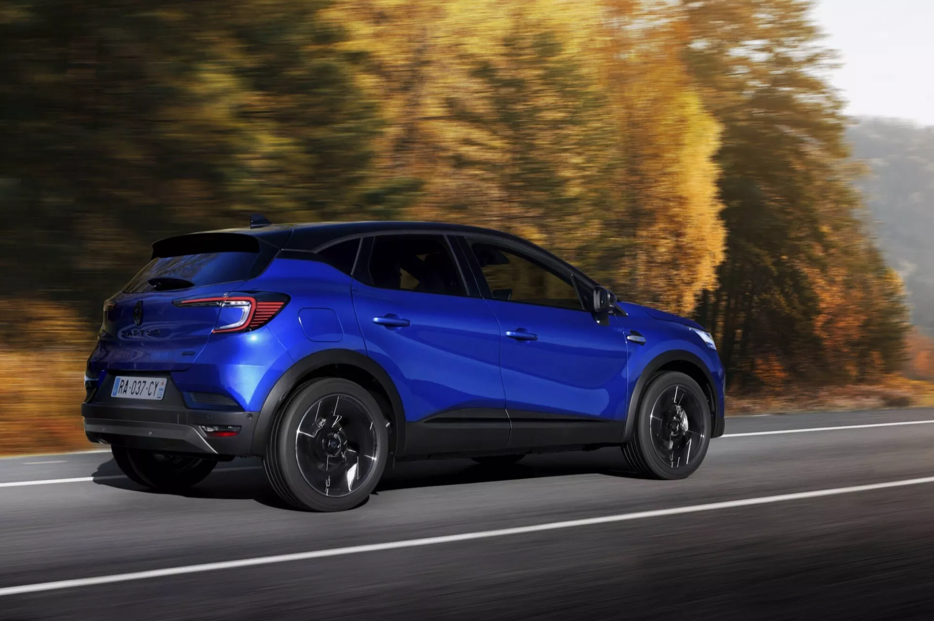 Veículo utilitário esportivo azul Renault Captur 2024 (II-facelift) em movimento dinâmico, vista de perfil destacando elementos de design lateral e traseiro em uma estrada com fundo de outono