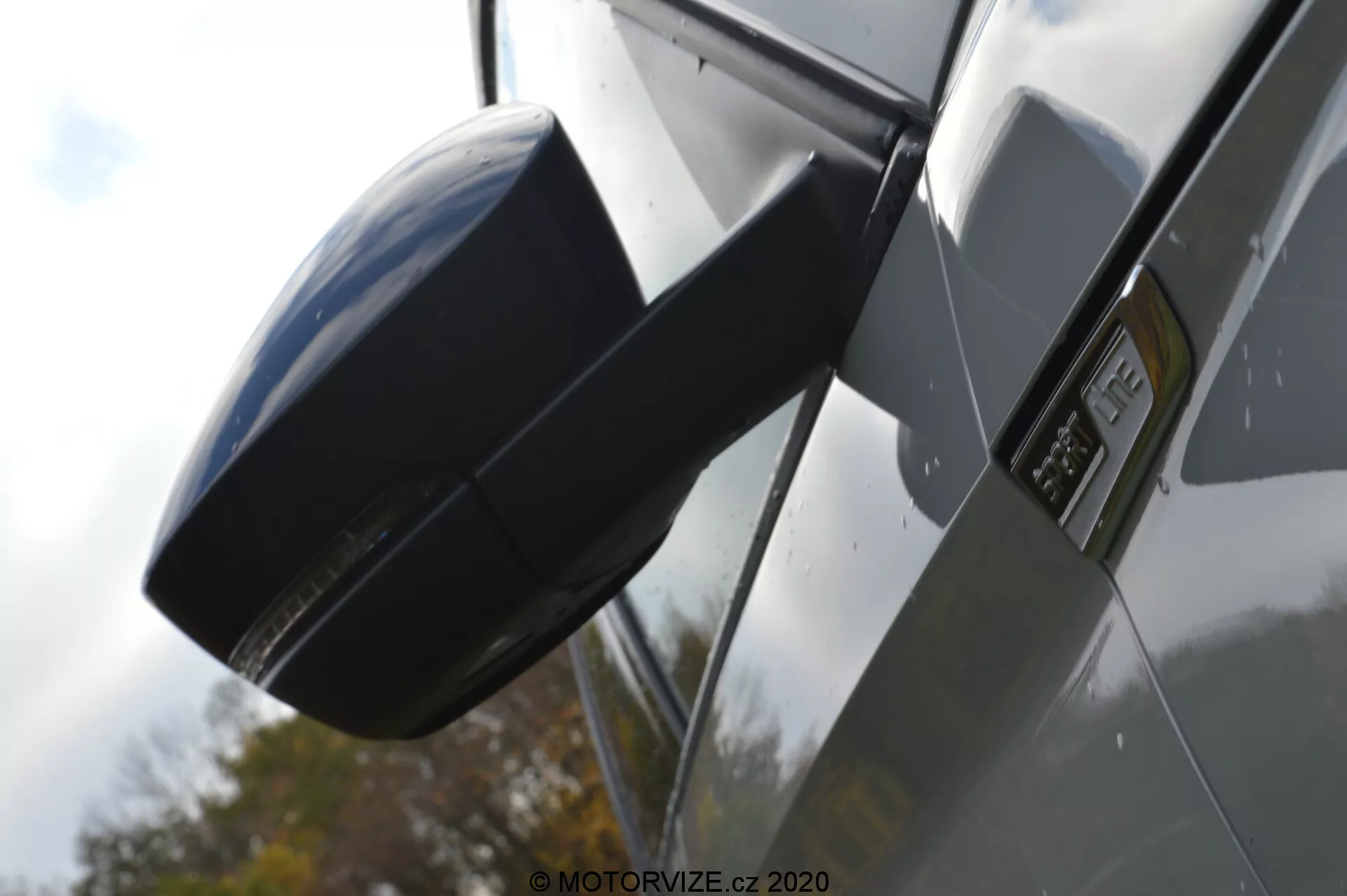 Vista lateral de close-up de um Skoda Kodiaq 2017 focando no espelho retrovisor externo e área do para-lama. A imagem é tirada de um ângulo baixo, enfatizando o perfil lateral do carro, com uma carcaça do espelho lateral moderna e aerodinâmica combinando com a cor do veículo, um emblema proeminente no para-lama indicando um modelo ou nível de acabamento específico, painéis de carroceria metálicos brilhantes e reflexos que sugerem um ambiente externo com um carro de cor escura.