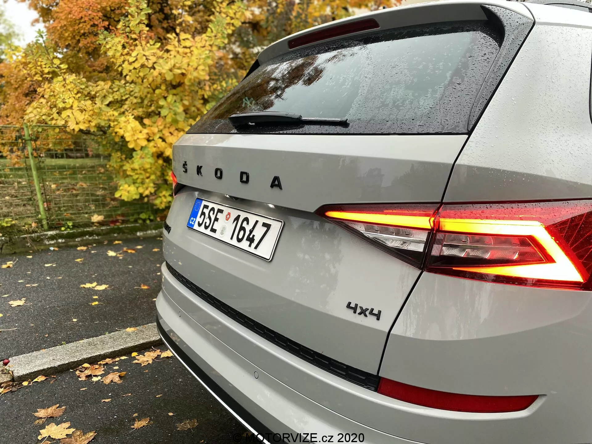 Vista traseira de um veículo Škoda prateado mostrando luzes traseiras de LED em forma de C iluminadas, emblema e marca Škoda, com emblema 4x4, capturada do lado traseiro direito em um pavimento molhado entre folhas de outono e árvores.