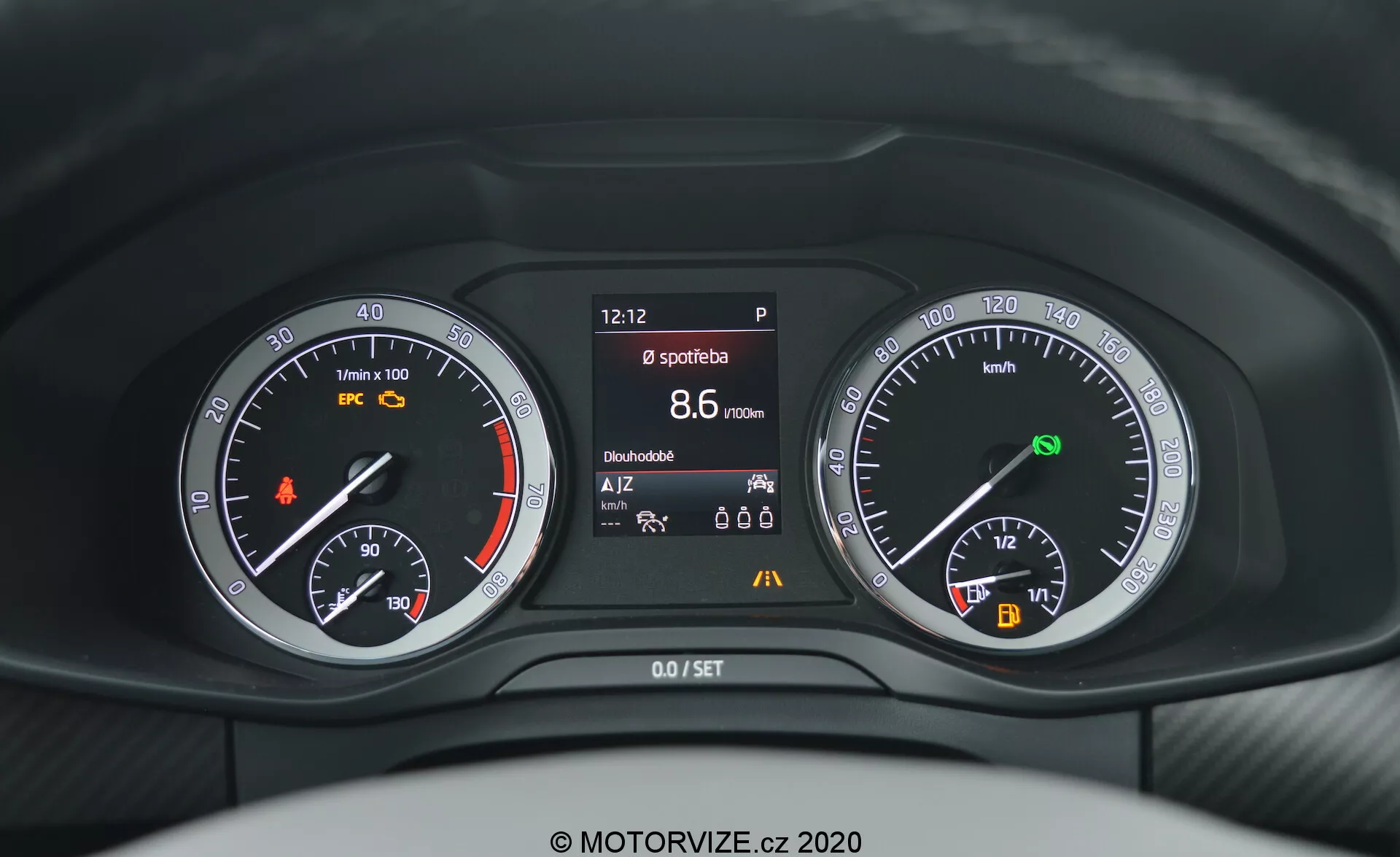Close-up da perspectiva do condutor do painel de instrumentos do Skoda Kodiaq 2017 mostrando uma combinação de medidores analógicos e digitais por detrás do volante. O painel inclui um tacômetro com uma luz de aviso laranja, uma tela central exibindo várias informações do veículo, como hora, status do freio de estacionamento, modo esportivo, alcance e temperatura, e um velocímetro com indicador de combustível e de start/stop automático.