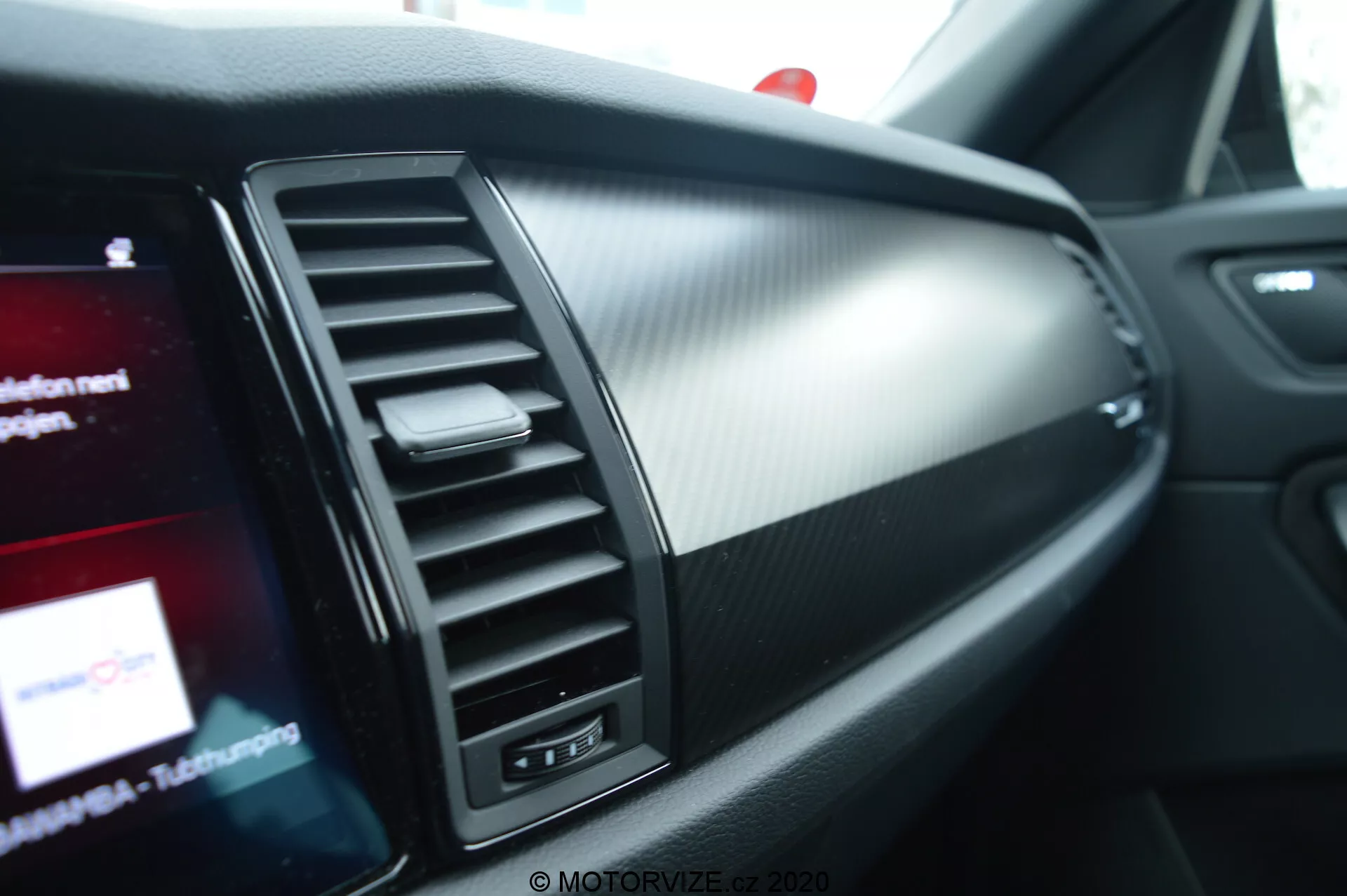 Vista aproximada do interior do Skoda Kodiaq 2017, destacando o lado do passageiro do painel. As características incluem uma saída de ar condicionado com aletas ajustáveis verticalmente, uma seção decorativa e uma tela de infotainment parcialmente visível com gráficos coloridos. Os materiais do painel combinam metal escovado e plástico texturizado que se assemelha a fibra de carbono, enfatizando um design moderno e esportivo.