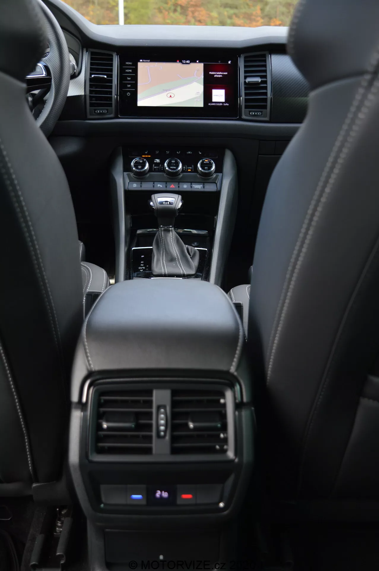 Interior do carro Skoda Kodiaq 2017, mostrando o painel frontal a partir da perspectiva do assento traseiro. A imagem destaca os assentos de couro, console central com câmbio automático, botões de controle, sistema de infotainment com tela sensível ao toque exibindo um mapa de navegação, saídas de ventilação do controle de clima e uma visão parcial do painel de instrumentos mostrando detalhes operacionais críticos.