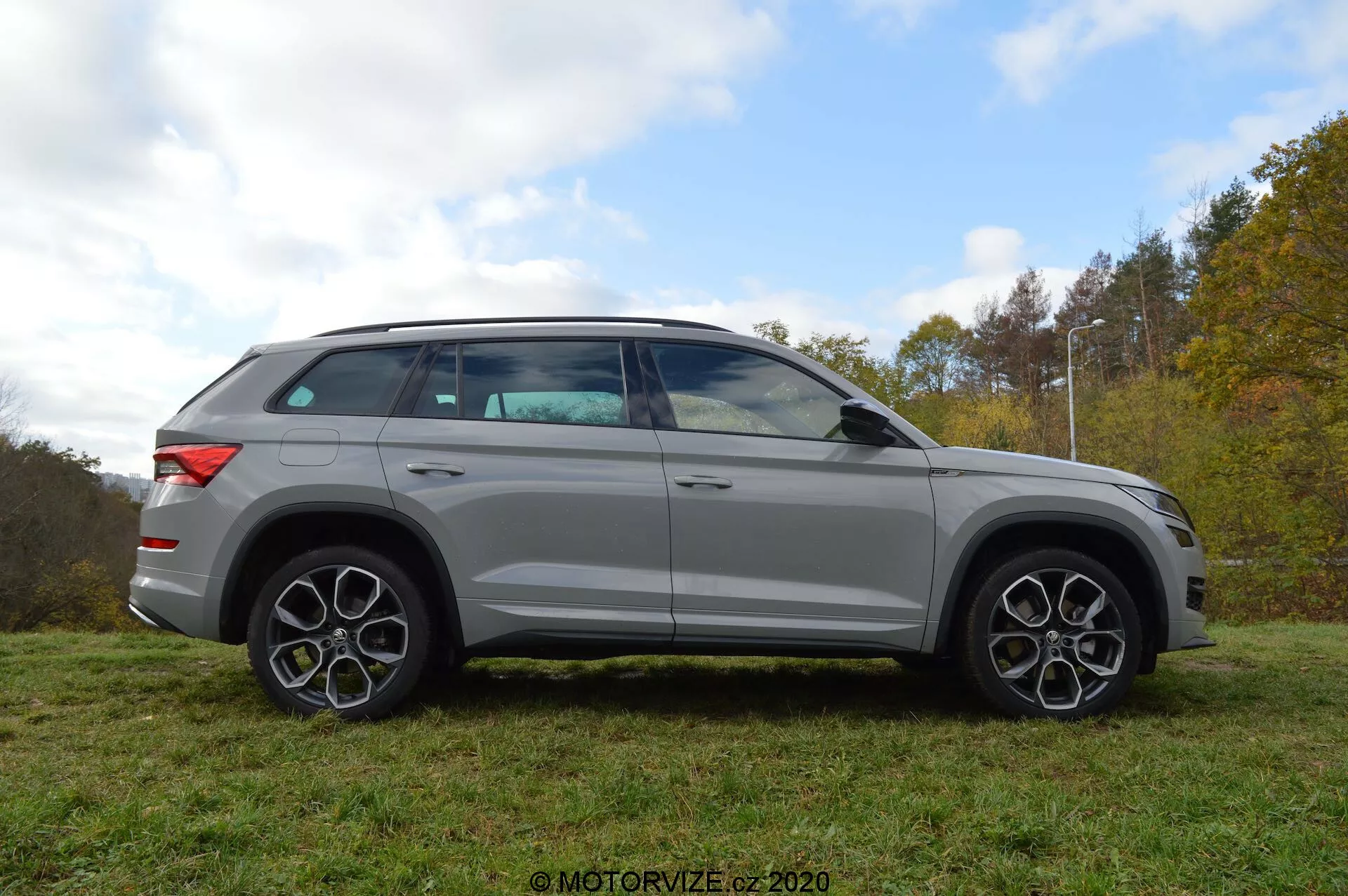 Vista de perfil completo de um SUV Skoda Kodiaq 2017 prateado estacionado na grama com um fundo de árvores e céu nublado. O design do veículo apresenta linhas arrojadas, rodas de liga grandes e jantes distintas com múltiplos raios. Os espelhos, que possuem indicadores de direção integrados, estão montados logo abaixo da linha das janelas. As janelas são ligeiramente escuras e a linha do teto inclina-se suavemente para trás, indicando um perfil esportivo, semelhante a um cupê. O SUV também está equipado com um sistema de trilhos no teto.