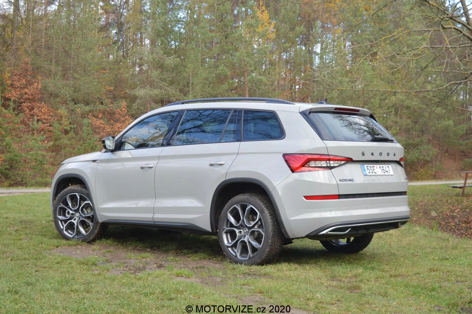 Imagem de um Škoda Kodiaq 2017 capturada de um ângulo traseiro três-quartos, mostrando a cor prata/cinza claro do veículo, rodas de liga leve de múltiplos raios, vidros traseiros escurecidos, teto solar panorâmico e design distintivo das luzes traseiras. O carro está estacionado ao ar livre com um fundo de árvores e grama, indicando um ambiente natural. No porta-malas traseiro são visíveis os emblemas Škoda e o distintivo de tração '4x4'.