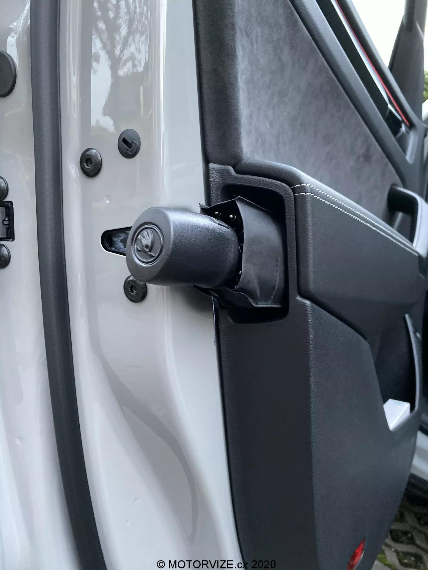 Vista aproximada do robusto mecanismo da dobradiça da porta e da área circundante de um Škoda Kodiaq 2017, com um componente de mola visível para uso pesado. O veículo branco contrasta com os componentes da dobradiça em preto, incluindo um mecanismo de fechadura tradicional com orifício para chave acima da dobradiça, fixado com parafusos de cabeça hexagonal para durabilidade. O plástico texturizado do painel interno da porta e os pontos de montagem seguros reforçam o design utilitário. Elementos de fundo desfocados destacam a dobradiça e o mecanismo de fechamento.