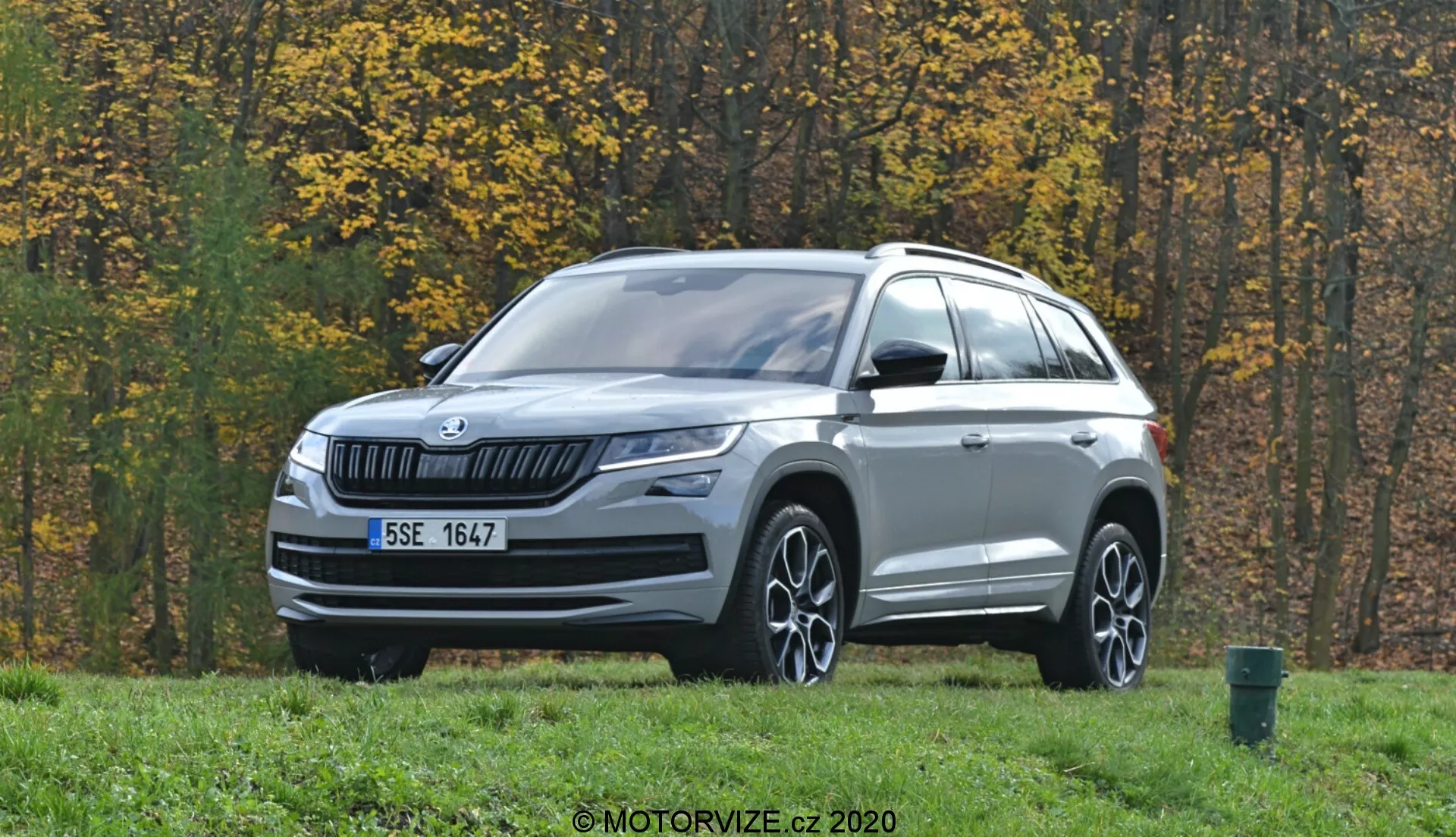 A imagem mostra um SUV Skoda Kodiaq 2017 estacionado em uma área gramada com árvores de outono ao fundo. Fotografado a partir de uma visão frontal de três quartos, o veículo apresenta uma grade frontal com lâminas horizontais e o logotipo da Skoda no centro. Os elegantes faróis LED se estendem para os lados, acompanhados por faróis de neblina na parte inferior do para-choque. O capô tem linhas sutis e um acabamento prateado adorna a parte inferior do para-choque dianteiro. Rodas de liga leve com design multirraios, espelhos laterais com indicadores de direção integrados, racks no teto e linhas esculpidas destacam o perfil. Sensores ou câmeras na parte inferior do para-choque sugerem avançados sistemas de assistência ao motorista (ADAS). O ambiente externo inclui uma estrada asfaltada e uma lixeira verde na borda direita da imagem.