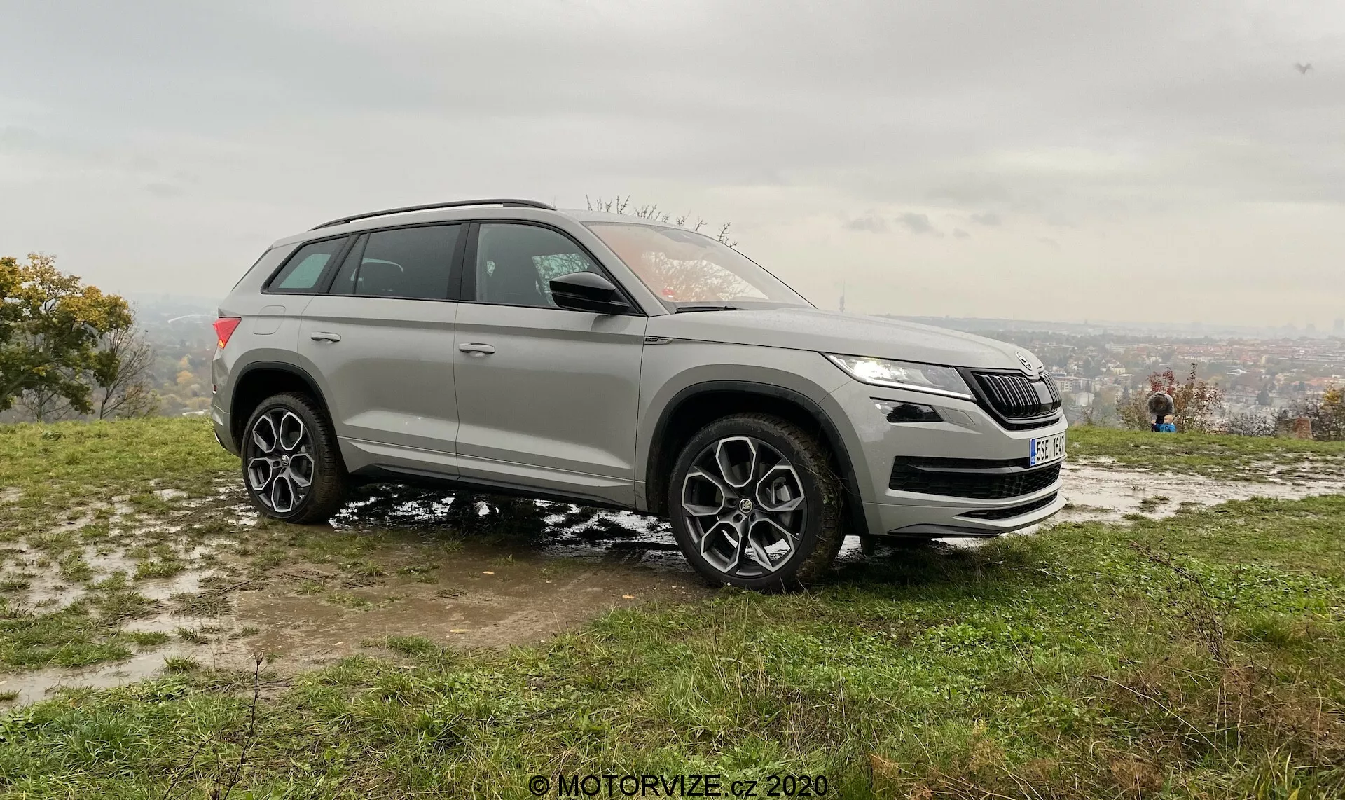 Imagem de perfil lateral de um SUV moderno tirada em ângulo oblíquo que mostra tanto os elementos de design frontal quanto lateral, ajustada contra um fundo natural de um ponto de vista elevado. A imagem destaca a grade frontal, faróis, luzes de neblina e logotipo distintivo do carro. Também apresenta elevada altura do solo, grandes rodas de liga leve com design de múltiplos raios, arcos de rodas proeminentes, linha de ombro alta, espelhos laterais, trilhos de teto e janelas escurecidas. O design esportivo e as tecnologias avançadas potenciais do veículo são acentuados nesta imagem diurna estacionária com manchas de neve, indicando uma estação mais fria.