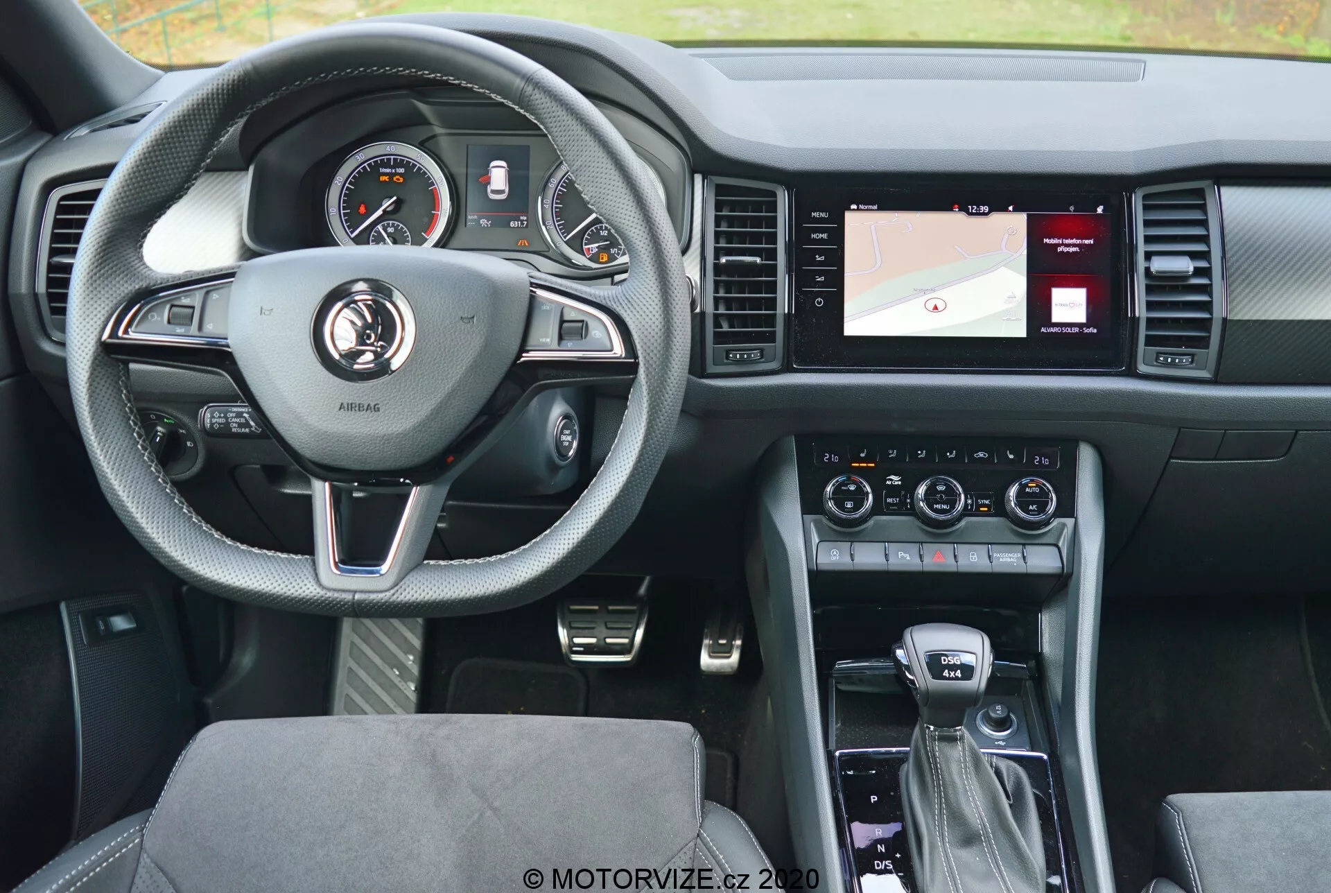 Vista interior de um Skoda Kodiaq 2017 a partir do banco traseiro, destacando o volante de três raios revestido em couro com detalhes prateados e botões de controle para o piloto automático, áudio e comunicação mãos-livres. O painel de instrumentos atrás do volante apresenta mostradores analógicos e um display digital. A consola central à direita inclui um ecrã com um mapa de navegação, controlos de climatização e configurações de áudio. A alavanca de mudanças sugere uma transmissão automática, com controladores adicionais para o sistema de infotainment e modos de condução. O design moderno visa um equilíbrio entre conforto e tecnologia, com assentos estofados em tecido com padrões e um layout bem iluminado e de fácil utilização.