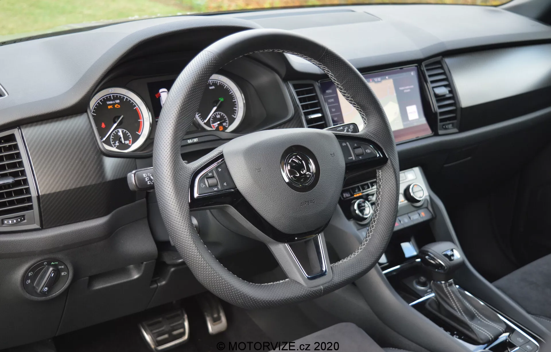 Vista interna de um Skoda Kodiaq 2017 focada na perspectiva do motorista do painel de instrumentos. O volante de três raios, revestido em material que lembra couro com costuras contrastantes, possui controles embutidos para áudio, comunicação viva-voz e configurações de controle de cruzeiro. Os medidores analógicos, com velocímetro à esquerda e tacômetro à direita, são complementados por um display digital no meio. A tela sensível ao toque de infotainment, ladeada por aberturas de ar, controles de clima e alavanca de câmbio são visíveis, juntamente com materiais texturizados e acentos metálicos escovados.