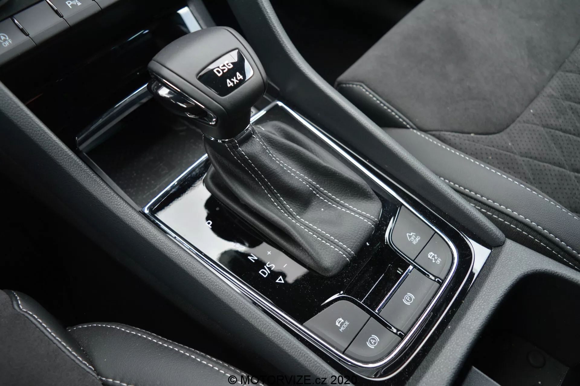 Vista aproximada da alavanca de câmbio automática e do console central preto brilhante do Skoda Kodiaq 2017, com modos de condução, travão de estacionamento eletrônico e botão de auto-hold; destaca as costuras em couro na manopla com modo esportivo.