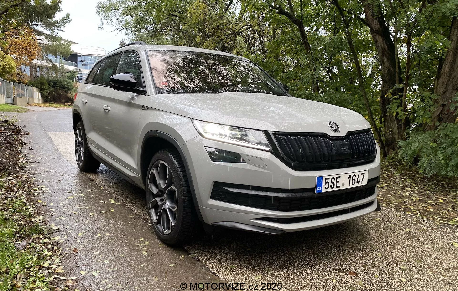 Skoda Kodiaq 2017 SUV moderno de uma marca europeia, ângulo de três quartos frontal mostrando o perfil frontal e lateral até o meio da porta traseira, estacionado ao lado de um caminho cercado por grama e árvores em um ambiente urbano ou suburbano. Características incluem grade dianteira característica com lâminas verticais, faróis de LED elegantes, luzes de nevoeiro no para-choque, grandes entradas de ar no para-choque dianteiro, linhas de contorno no capô para aerodinâmica, arcos de roda proeminentes com rodas de liga leve de design multirraio, espelhos laterais com indicadores de seta integrados, molduras protetoras na parte inferior e pintura geral limpa e bem preservada.