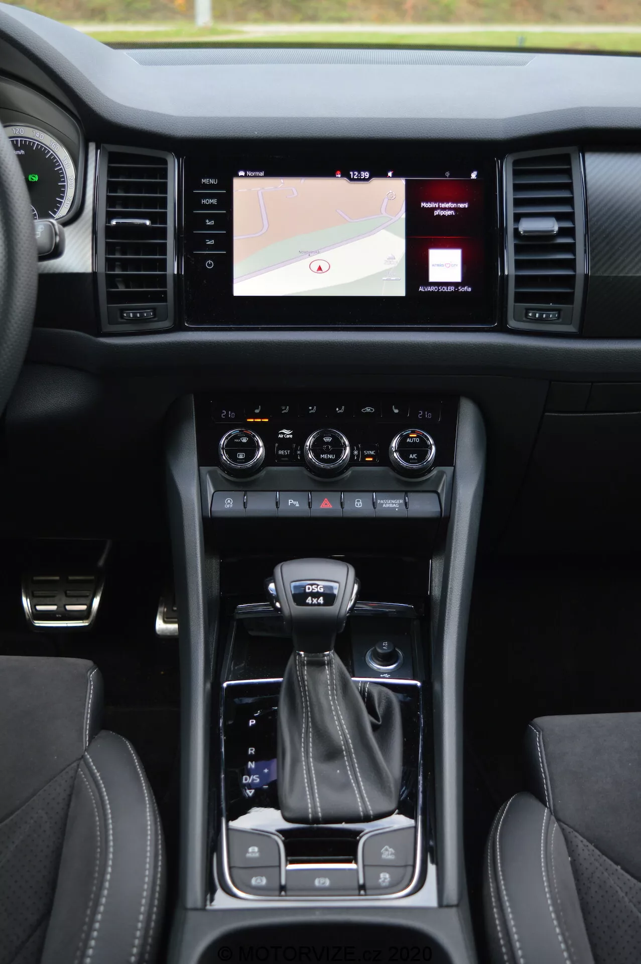 Vista interna de um Skoda Kodiaq 2017 mostrando o console central, sistema de infoentretenimento com tela sensível ao toque mostrando um mapa, botões duplos de controle climático, alavanca de câmbio, seletor de modo de condução, porta-copos, bancos de couro com costura contrastante e botão de partida sem chave