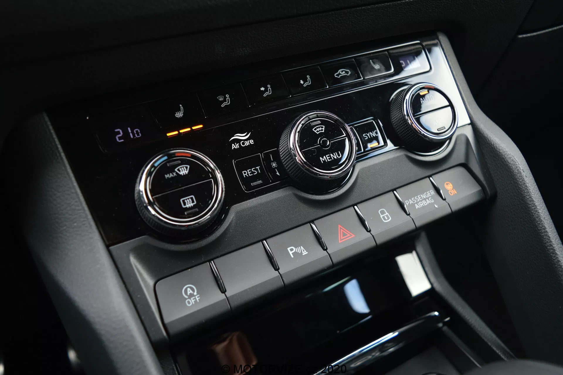 Close-up da consola central do Škoda Kodiaq 2017, destacando o sistema de controlo climático com dois grandes botões de ajuste de temperatura e um visor digital definido para 21 graus Celsius. A imagem também apresenta botões para ar condicionado, recirculação, descongeladores de vidros e um botão 'MENU'. Abaixo estão os interruptores basculantes para sensores de estacionamento, desativação do start/stop automático e luzes de emergência.