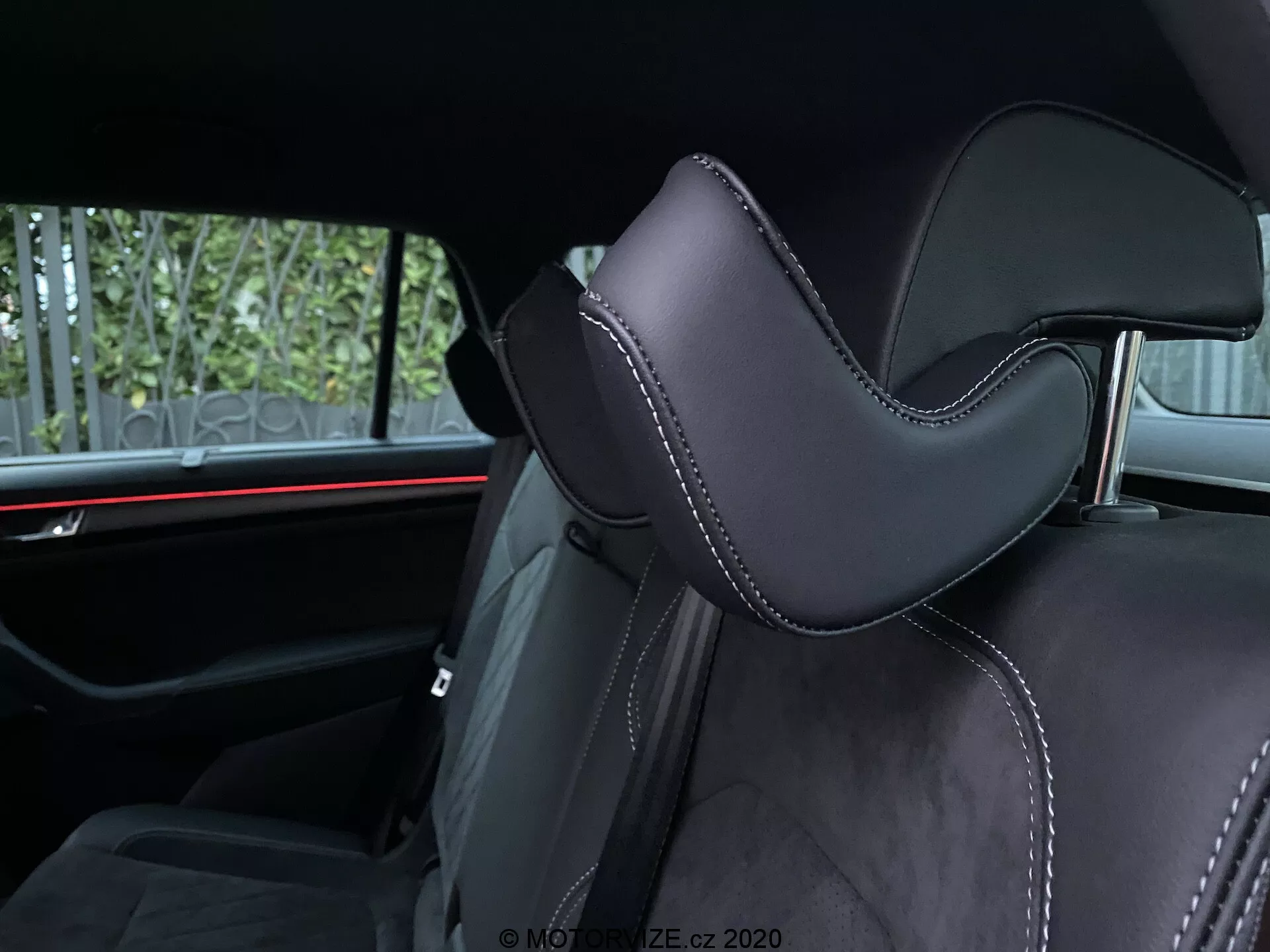 Vista interior de um Skoda Kodiaq 2017 focada em um assento de carro. A captura angular enfatiza a parte do apoio de cabeça, estofada em material preto, provavelmente couro ou sintético de alta qualidade, com contornos distintos para conforto. Costuras precisas com fio contrastante adicionam detalhes luxuosos. Ao fundo, uma iluminação ambiente horizontal vermelha destaca os elementos de design automotivo moderno. As comodidades visíveis para os passageiros traseiros incluem cintos de segurança e possivelmente um apoio de braço central, indicando a segunda fila de assentos.