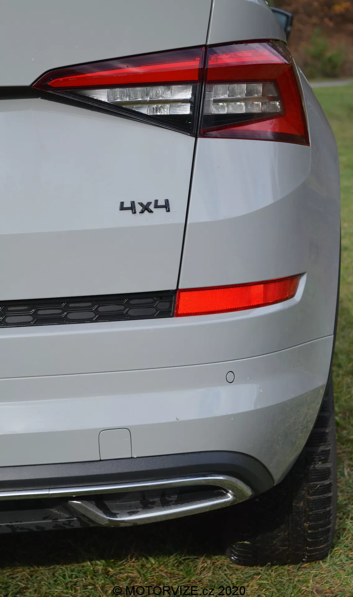Vista de perto do quarto traseiro esquerdo de um Skoda Kodiaq 2017. A imagem foca na lanterna traseira, com luzes LED modernas de design angular e nítido, e divisões claras para diferentes funções de luz. O emblema '4X4' indica tração nas quatro rodas. O para-choques traseiro inclui refletores e sensores, provavelmente para assistência de estacionamento, e uma ponta de escape visível. A pintura metálica prateada e as linhas sutis da carroceria são mostradas, com um fundo de grama e árvores, sugerindo um ambiente suburbano ao ar livre. Marca d'água 'MOTORVIZE.cz 2020'.
