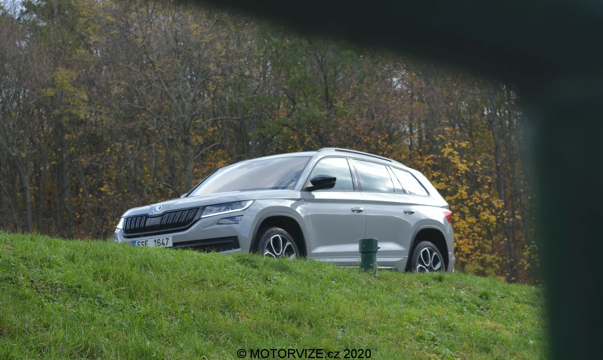 Vista de perfil lateral de um SUV Skoda Kodiaq 2017 branco estacionado em uma colina gramada, voltado para a esquerda, com jantes de liga e pintura metálica refletindo a luz do sol. O ambiente inclui folhagem e árvores, sugerindo uma localização rural ou em parque. Visto de um ângulo baixo através de um primeiro plano borrado, o veículo apresenta elementos de design moderno, como faróis de LED, espelhos com sinalizadores integrados, vidros escurecidos, trilhos de teto e uma antena de barbatana de tubarão.