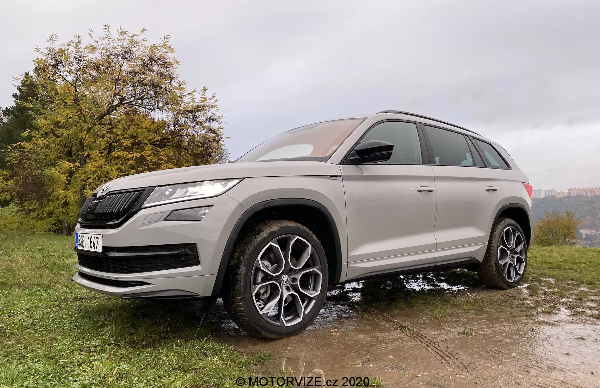 Vista de três quartos da frente de um SUV Škoda Kodiaq 2017 tirada de um ângulo ligeiramente baixo, enfatizando a sua altura e postura. O veículo está estacionado em uma superfície irregular de grama e lama, destacando suas capacidades off-road. O Škoda Kodiaq possui uma grade proeminente e faróis LED afiados. O design meticuloso inclui grandes rodas de liga leve com múltiplos raios, alta distância do solo e pneus para todas as estações. A carroceria é pintada em uma cor metálica neutra com vidros escurecidos, e os toques de cromo realçam o seu apelo premium. As funcionalidades modernas são notadas nos retrovisores laterais na cor da carroceria com indicadores de direção integrados e possivelmente sistemas de aviso de ponto cego.