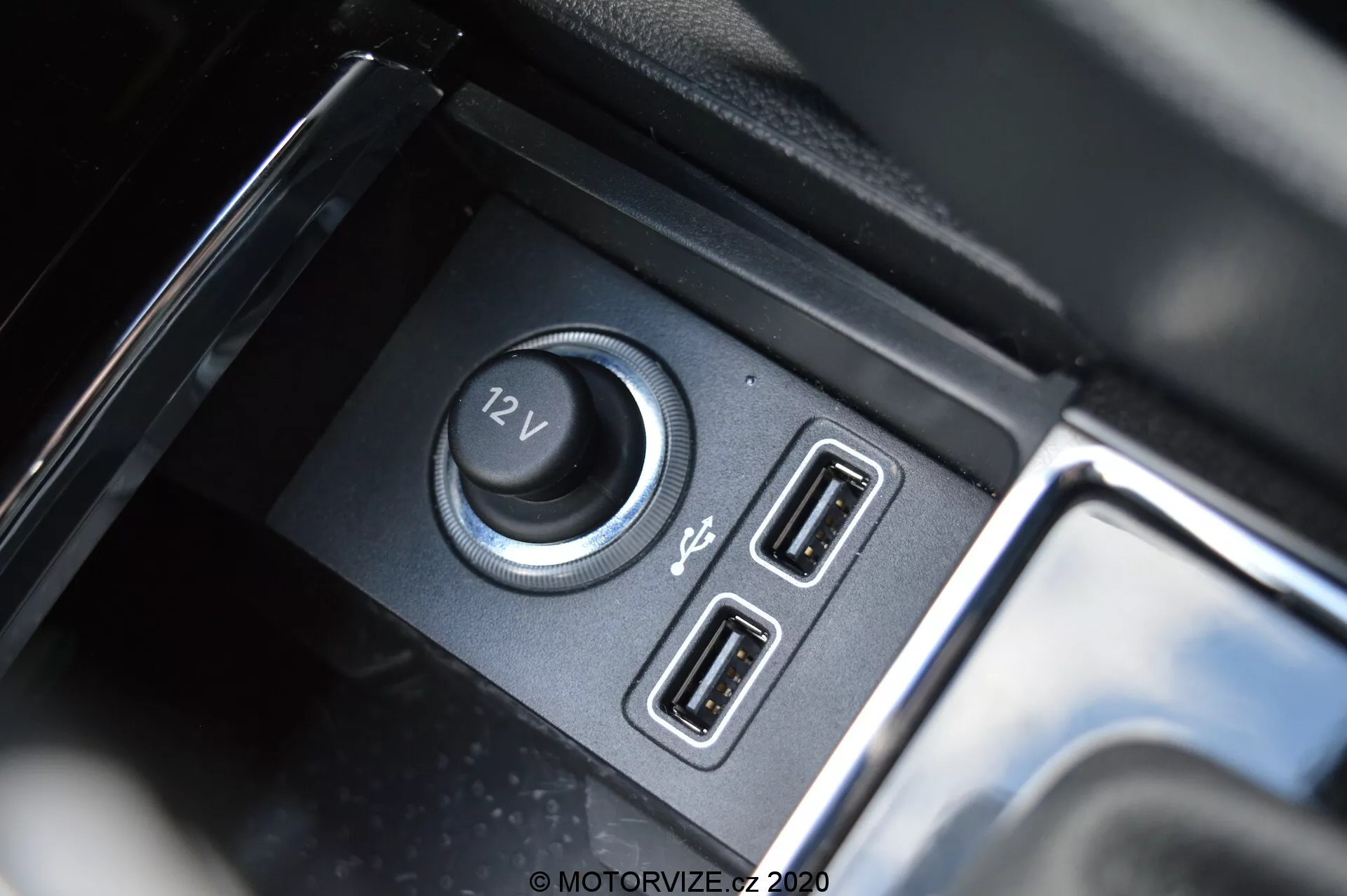 Close-up do interior do Škoda Kodiaq 2017, destacando as funções de alimentação e conectividade, incluindo uma tomada de 12V e portas USB duplas, com compartimento preto fosco e detalhes brilhantes e reflexivos.