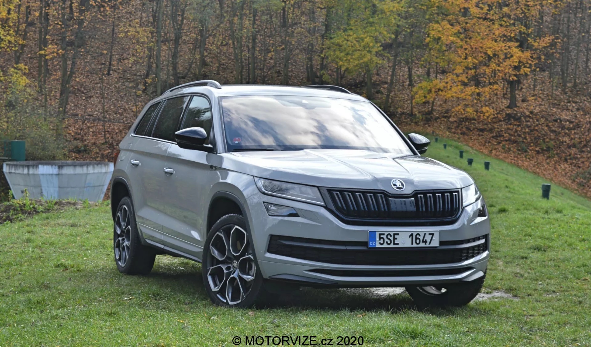 Uma visão detalhada do Škoda Kodiaq 2017, um SUV de cor prata, posicionado ao ar livre contra um cenário de outono de árvores. A imagem diurna captura o veículo de um ângulo de três quartos dianteiro, mostrando seu perfil frontal e lateral. Elementos de design distintivos são visíveis, incluindo o emblema da marca na grade frontal, faróis de LED, luzes de neblina, rodas de liga de múltiplos raios, trilhos de teto e espelhos laterais com sinais de pisca integrados. O carro está posicionado na grama perto de uma borda de concreto, indicando um ambiente semelhante a um parque.