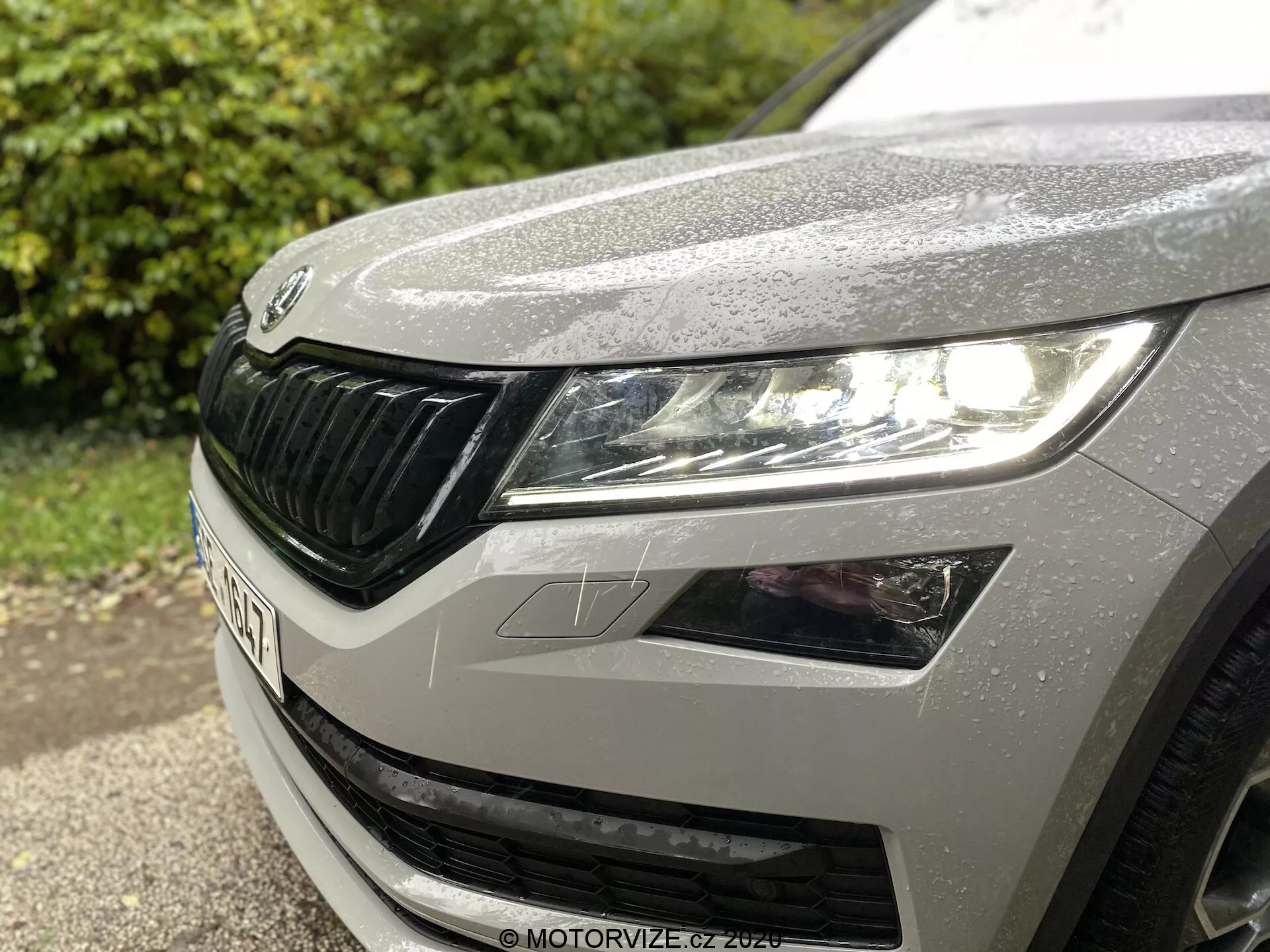 Vista aproximada do quarto dianteiro esquerdo de um Škoda Kodiaq 2017, mostrando o farol esquerdo com design moderno de LED, parte do capô com gotas de chuva e a grade distinta da Škoda com elementos horizontais.