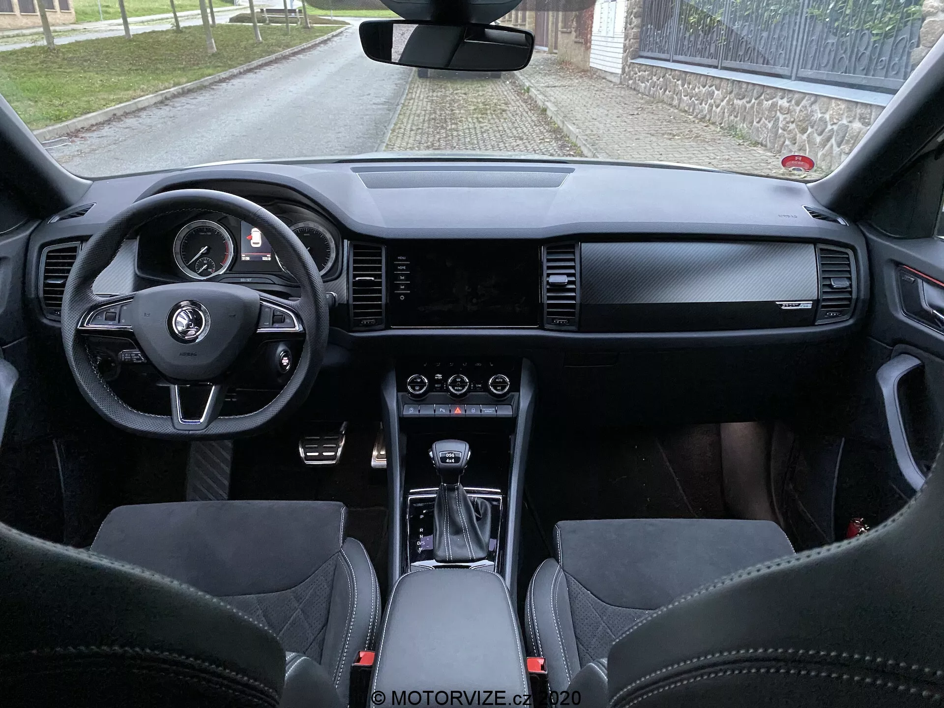 Vista interna do Skoda Kodiaq 2017 a partir do espaço do assento traseiro, mostrando o painel de instrumentos, volante com botões multifuncionais, cluster de instrumentos digital, tela ampla de infotainment, controles HVAC, seletor de marchas automático, materiais de couro premium e assentos esportivos com encostos de cabeça integrados.