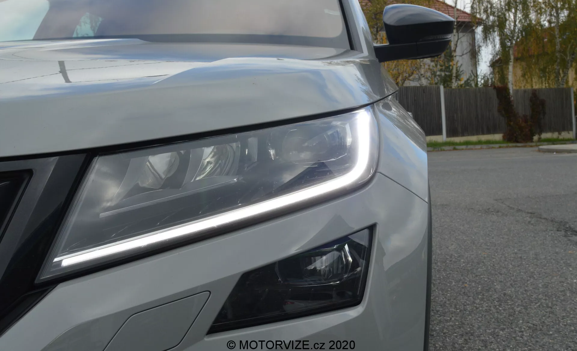 Close-up da frente esquerda de um Skoda Kodiaq 2017. A perspectiva de ângulo baixo destaca as distintas luzes de condução diurna em LED e os faróis principais detalhados que sugerem uma tecnologia de iluminação avançada. Luzes de nevoeiro com design moderno são visíveis abaixo do farol principal. A imagem também mostra parte da grade frontal, pintura metálica com linhas de carroçaria acentuadas e um espelho lateral com indicador de seta integrado. Há uma marca d'água 'MOTORVIZE.cz 2020' indicando a fonte da fotografia.