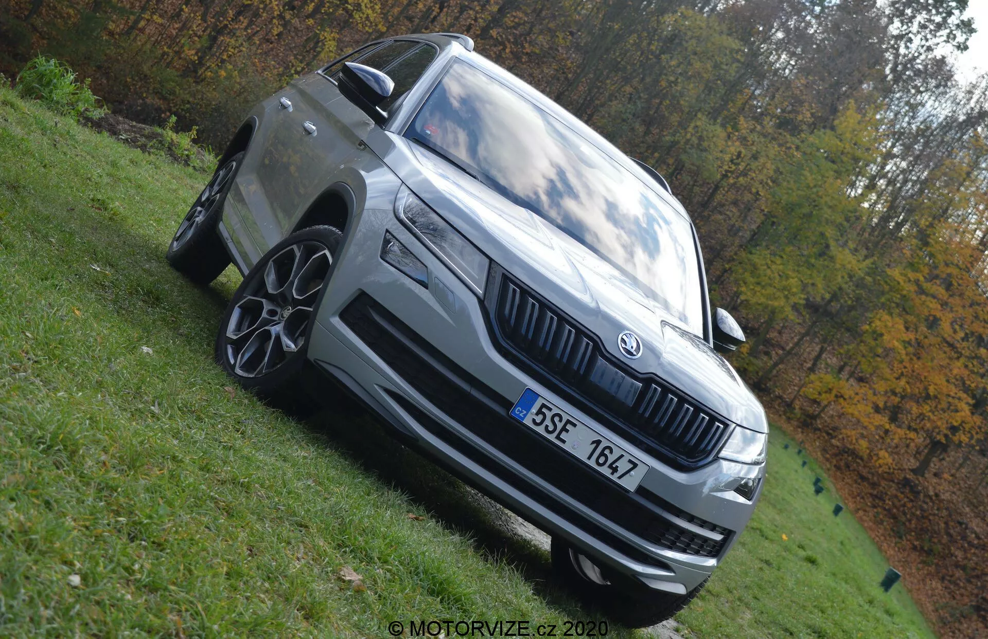 Fotografia em ângulo baixo do Skoda Kodiaq 2017 destacando sua postura dominante. O carro é capturado mostrando seu quarto frontal esquerdo com características notáveis como faróis de LED, grade proeminente com emblema Skoda, rodas de liga leve com vários raios, para-choque dianteiro com entradas de ar, espelho lateral com sinalizador integrado e linhas de design exterior distintas. O veículo é cinza claro ou prata e está estacionado em uma área gramada sob luz natural do dia. O fundo borrado de outono acentua a presença do veículo.