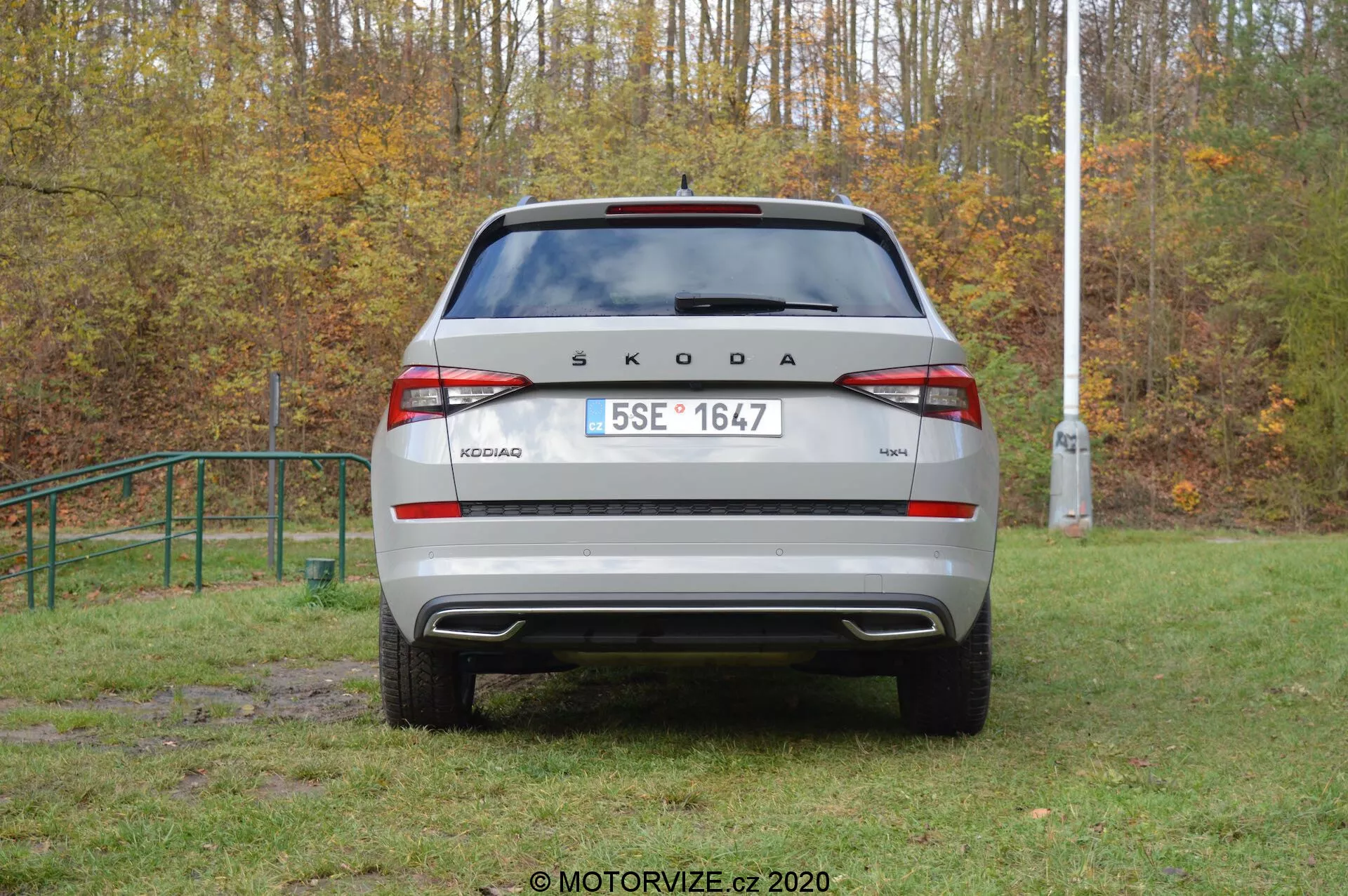 Imagem da traseira de um veículo Škoda Kodiaq 2017 em um ambiente externo. A marca 'ŠKODA' e o nome do modelo 'KODIAQ' estão exibidos de forma proeminente na tampa do porta-malas. Placa de licença no estilo europeu, luzes traseiras modernas com luz de neblina traseira integrada e luzes de ré, para-choque traseiro visível com sensores de estacionamento e para-brisa traseiro com limpador são claramente visíveis. Elementos refletores na parte inferior do para-choque aumentam a visibilidade. O carro parece estar estacionado em uma área gramada e ligeiramente lamacenta, sugerindo capacidade off-road.