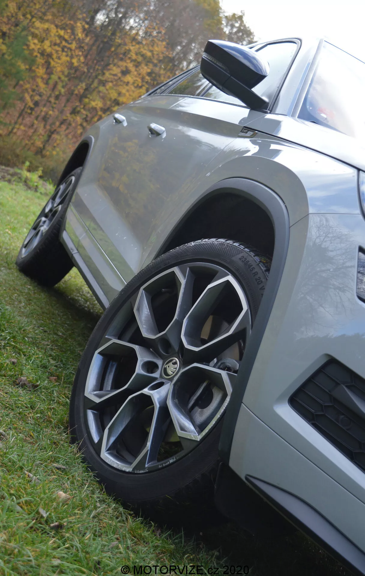 Close-up de baixo ângulo de um Škoda Kodiaq 2017 destacando a roda dianteira e a área do para-lama. O carro, estacionado na grama, apresenta uma roda de liga leve multifio, espelho lateral aerodinâmico com indicador integrado, painéis de carroceria angulares com acabamento em pintura metálica e uma ventilação no para-lama dianteiro, tudo em um fundo natural desfocado.
