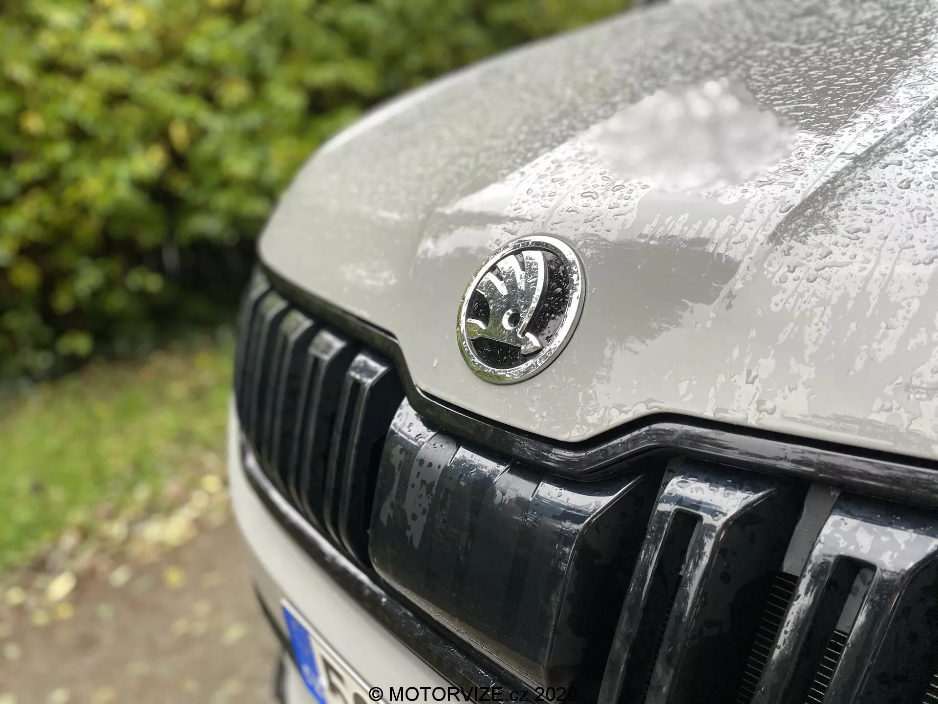 Imagem de close-up da seção frontal de um carro Skoda Kodiaq 2017, com foco principal na grade e no emblema do fabricante. O emblema circular está colocado no centro, exibindo detalhes delicados com gotas de água indicando chuva recente. O ângulo ligeiramente descentrado para cima captura o capô e as lamelas verticais refletoras da grade em foco nítido com uma profundidade de campo rasa.