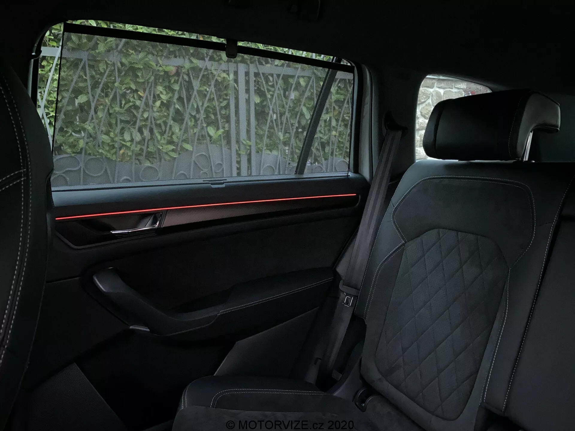 Vista interior do Skoda Kodiaq 2017 focando no compartimento traseiro dos passageiros, destacando características de luxo, como estofamento em couro com costura em padrão diamante nos assentos dianteiros e traseiros, tira de iluminação vermelha ambiental no painel da porta bem equipado com acabamento premium e uma tela de proteção solar parcialmente translúcida na janela. A ênfase está no conforto e nos elementos de design de alta qualidade.
