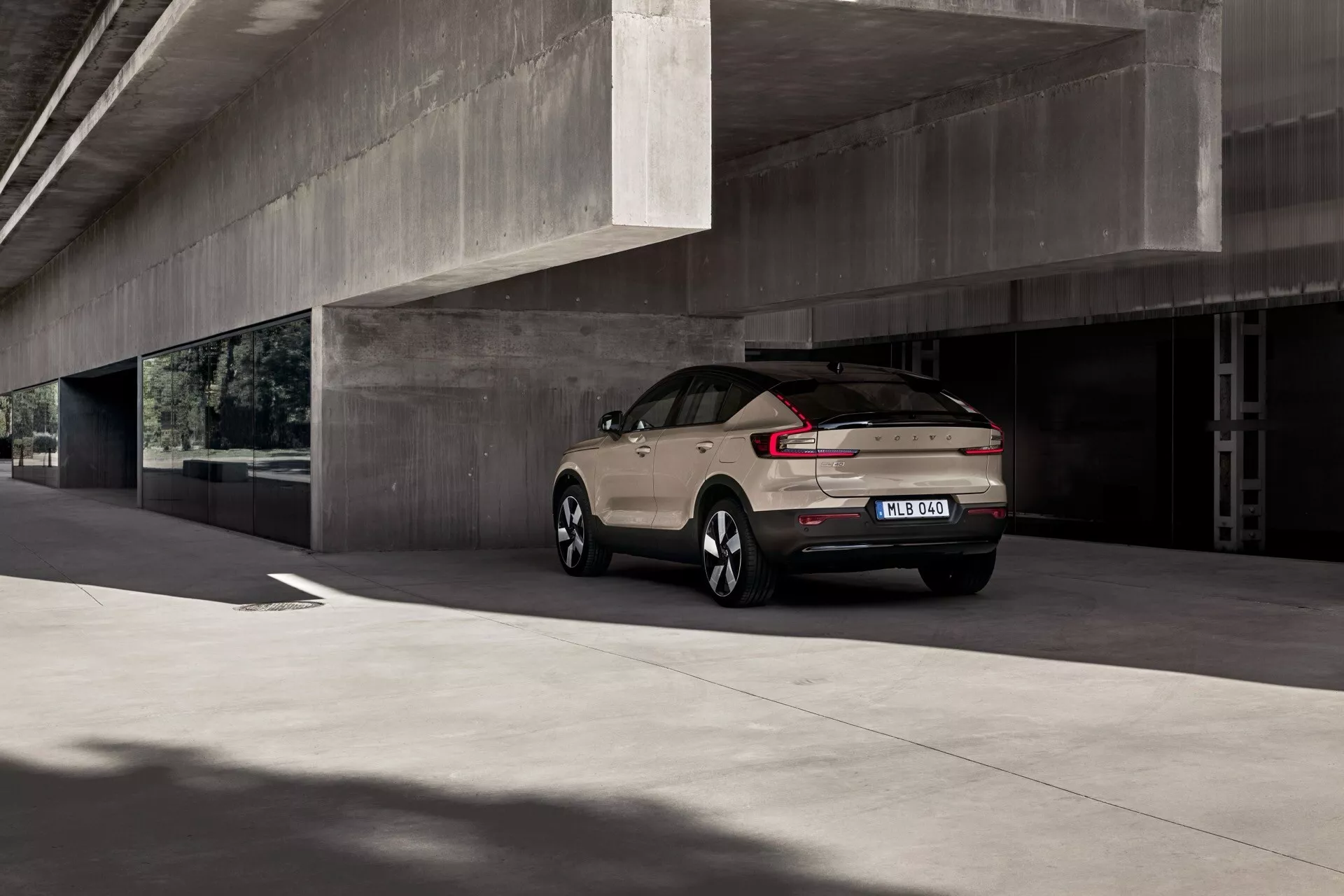 Volvo EC40 2024, 1ª geração, estacionado sob uma estrutura de concreto mostrando seu design moderno de SUV, com luzes traseiras aerodinâmicas, para-choque traseiro bem definido com sensores de estacionamento, rodas de liga leve multi-raios e antena de barbatana de tubarão, em uma cor neutra fosca, em um dia nublado.