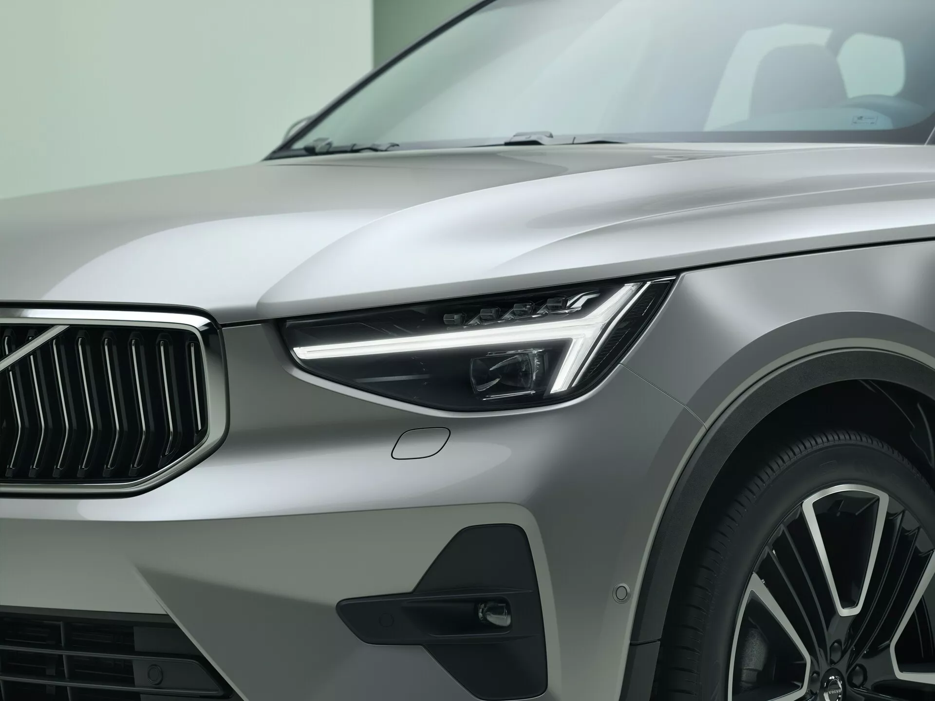 Close-up do quarto dianteiro esquerdo do modelo Volvo XC40 2022 de primeira geração facelift, exibindo farol de LED elegante com luzes diurnas, grade frontal proeminente com ripas verticais, pneu de baixo perfil de alto desempenho em roda de liga leve multirraios, pintura metálica e elemento adicional de design no para-lama, enfatizando luxo e design automotivo moderno.