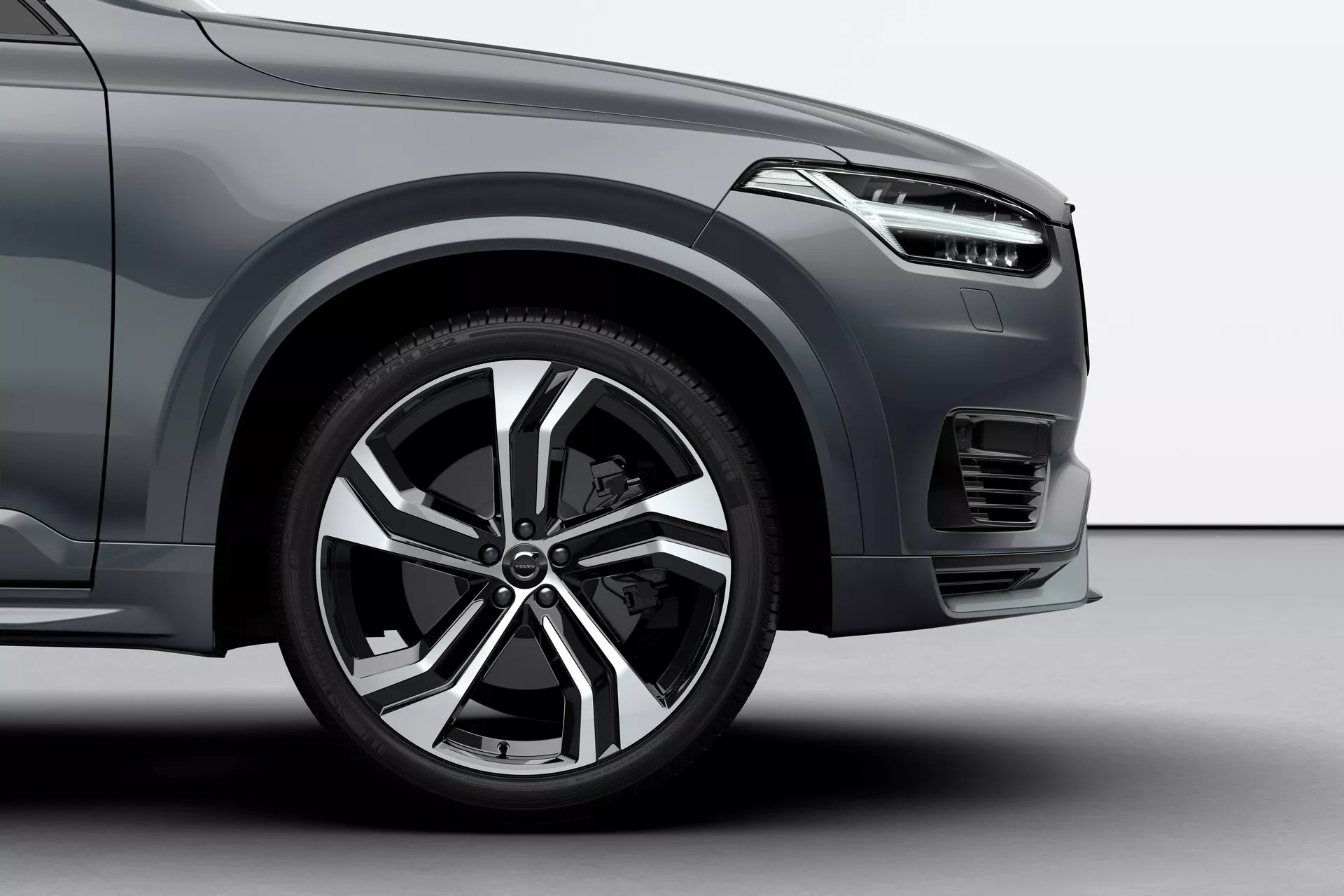 Close-up, visão em ângulo baixo da roda dianteira e seção inferior de um Volvo XC90 2015 (2ª geração). A foto mostra uma roda de liga leve com vários raios e acabamento em prata e preto, um pneu de perfil baixo focado no desempenho na estrada e um farol horizontal elegante parcialmente visível. A imagem também destaca um para-choque dianteiro contornado com uma pequena seção de grade, sugerindo um SUV moderno e premium. O carro ocupa a maior parte do quadro com um fundo neutro, provavelmente um estúdio.