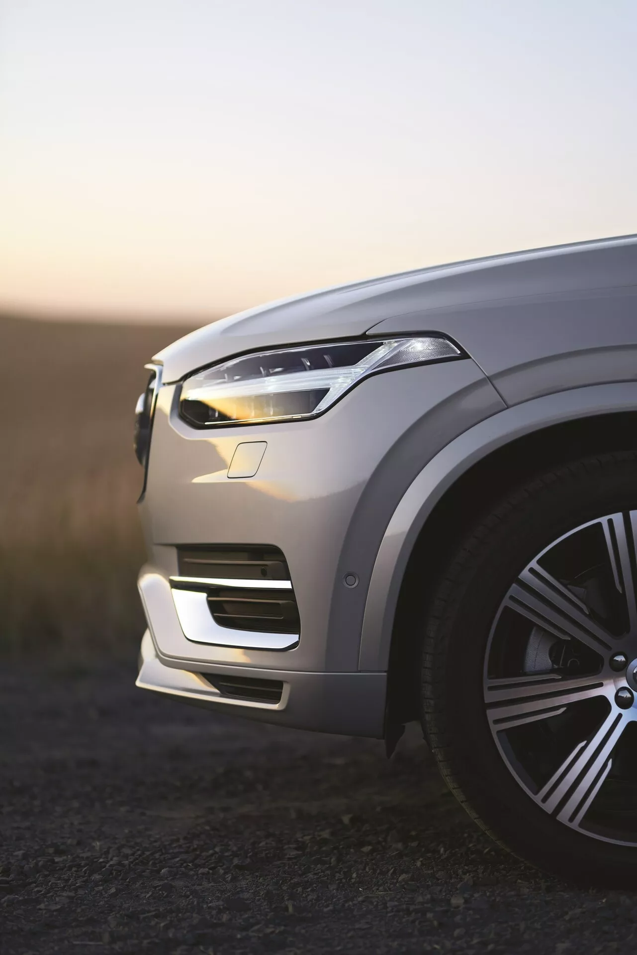 Vista do quarto dianteiro esquerdo do SUV de luxo Volvo XC90 de 2015, segunda geração, ao amanhecer, luzes diurnas de LED iluminadas, pintura metálica refletindo a luz ambiente, roda de liga grande e elegante, contornos intricados do capô, design elegante do farol de LED, para-choque dianteiro integrado com entradas de ar, possíveis sensores de assistência ao motorista visíveis, profundidade de campo rasa com fundo desfocado.