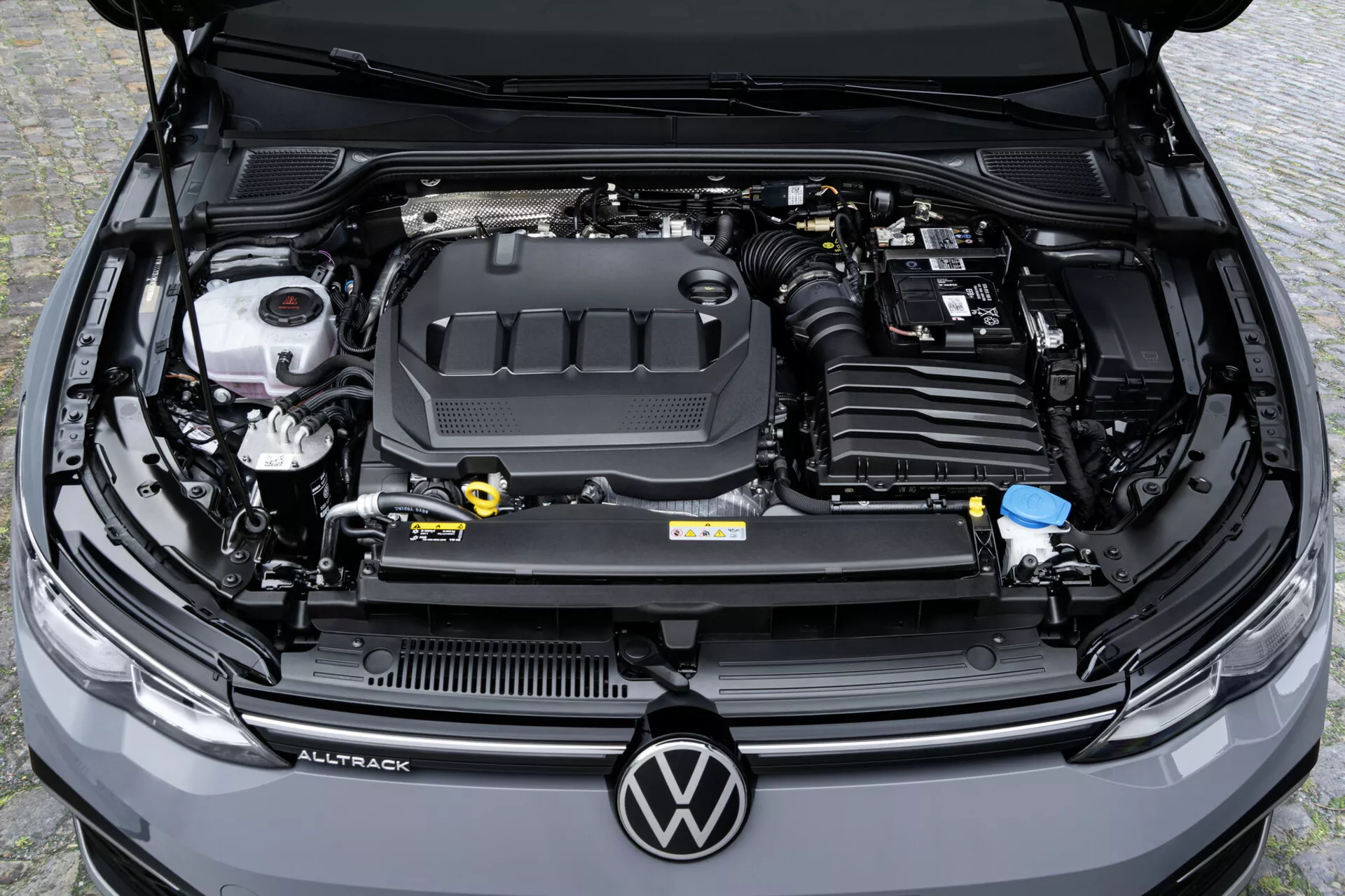Vista aérea de ângulo alto do compartimento do motor de um Volkswagen Golf Alltrack 2017 mostrando a capa do motor com logotipo, reservatórios de fluido e bateria do carro